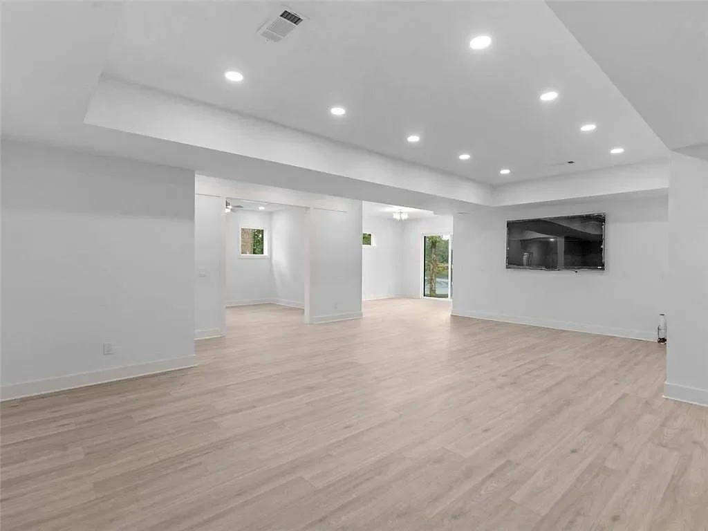 Empty room with white walls, light wood flooring, recessed ceiling lights, a flat-screen TV mounted on the wall, and multiple windows letting in natural light.