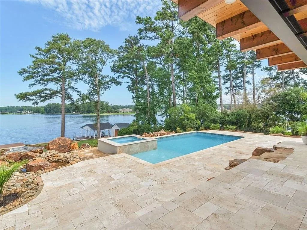 View of a backyard with a swimming pool and hot tub overlooking a lake with trees and a dock with small structures.
