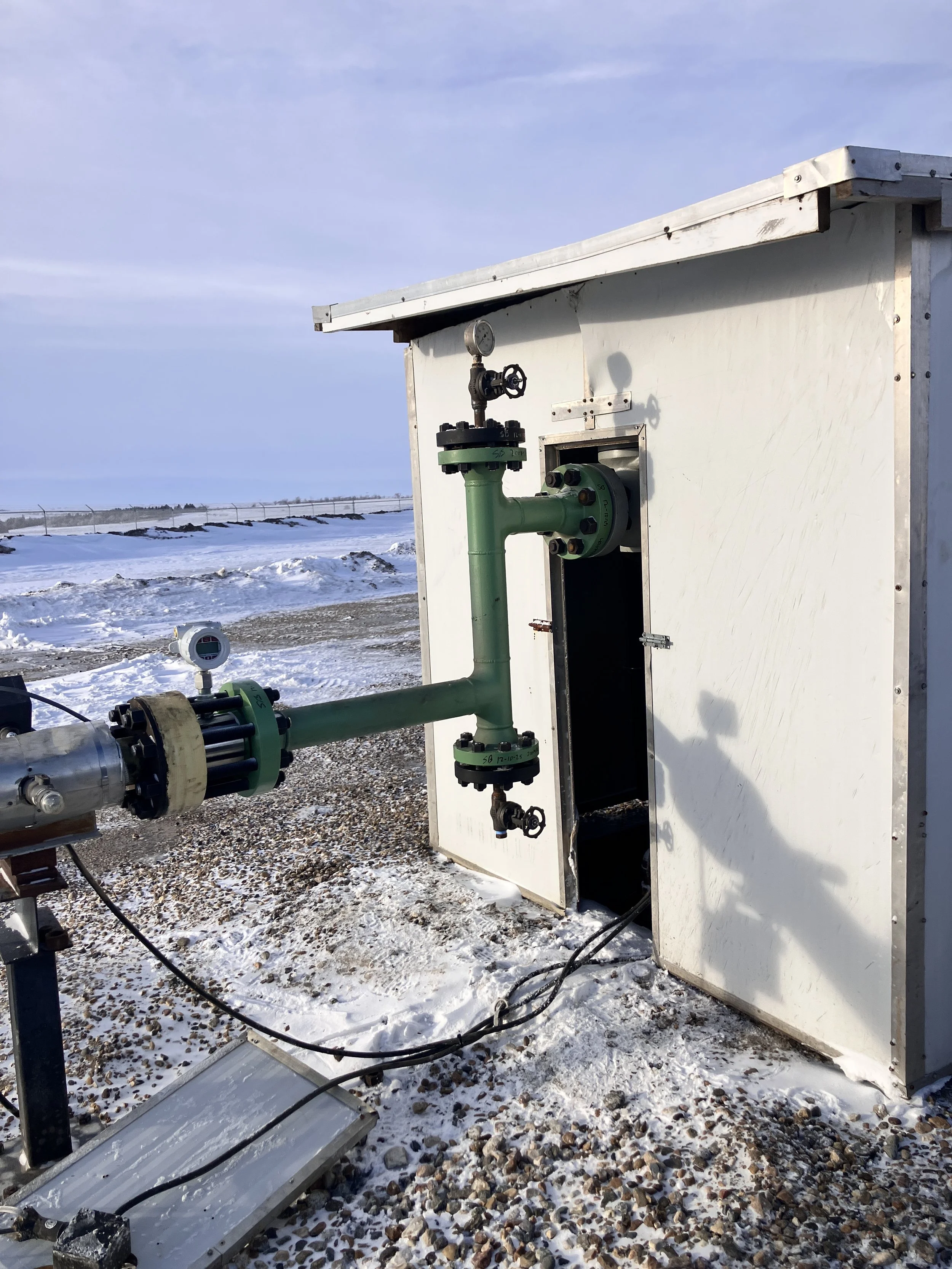 An outdoor scene with a white rectangular box-like structure containing green piping and gauges, situated on gravel and snow, with a fence and cloudy sky in the background.