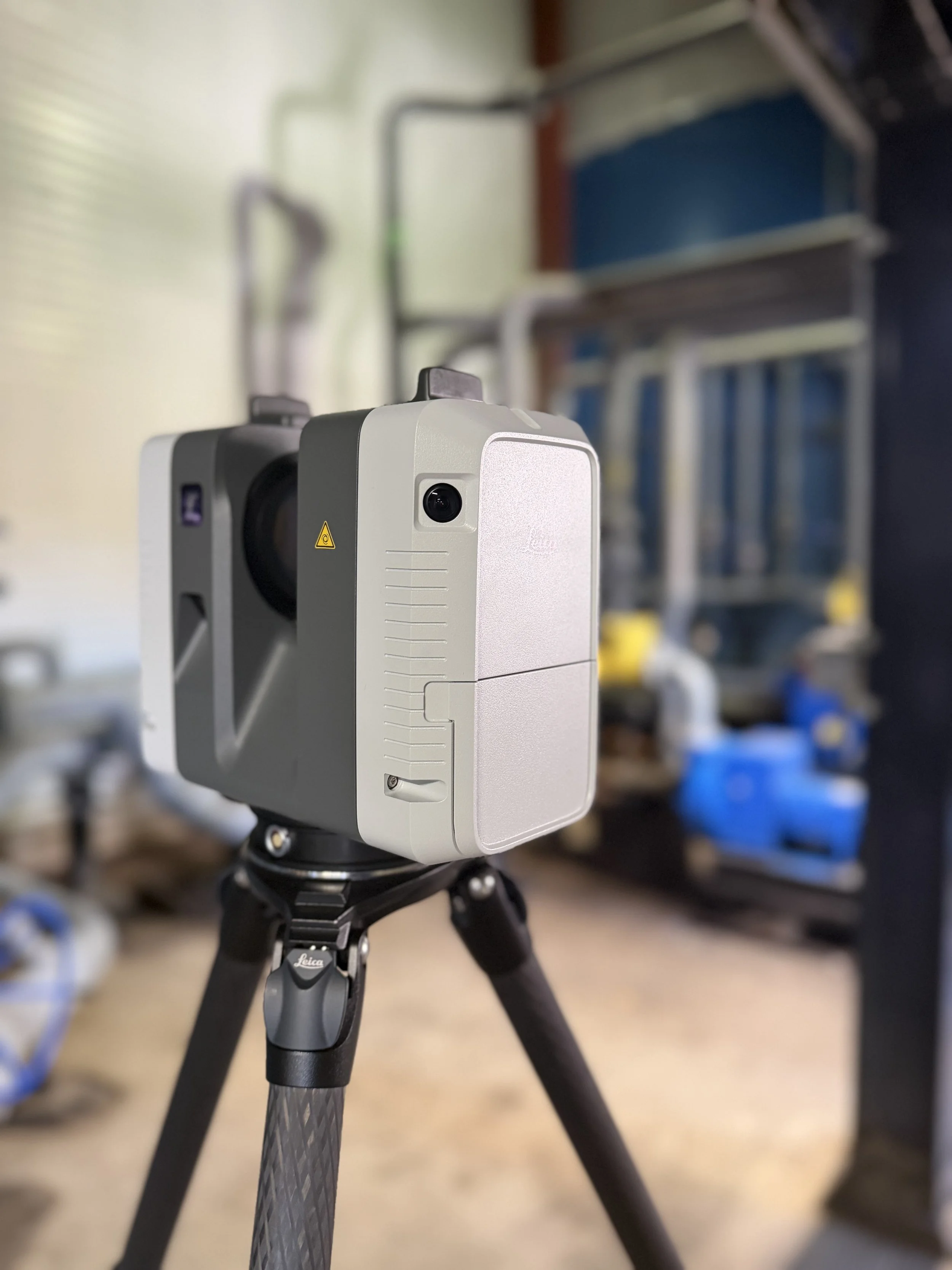 A Leica laser scanner mounted on a tripod, with a blurred background of equipment and shelves in a workshop or laboratory setting.