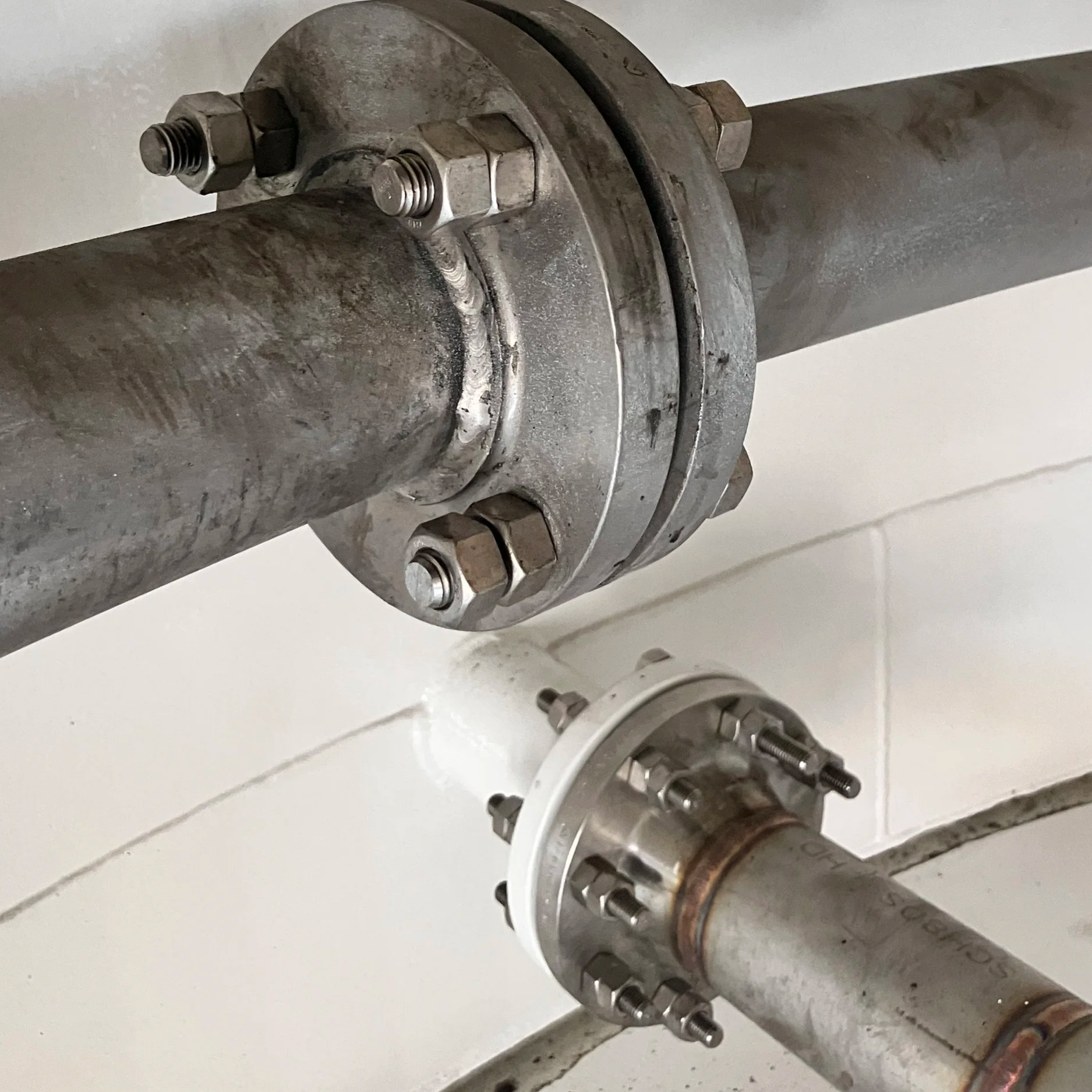 Close-up view of a metal pipe with a flange connection, showing bolts and nuts, with some rust and wear on the metal surfaces, on a white background.
