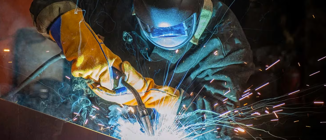 A person welding metal, wearing protective gloves and a welding helmet, sparks flying from the welding process.