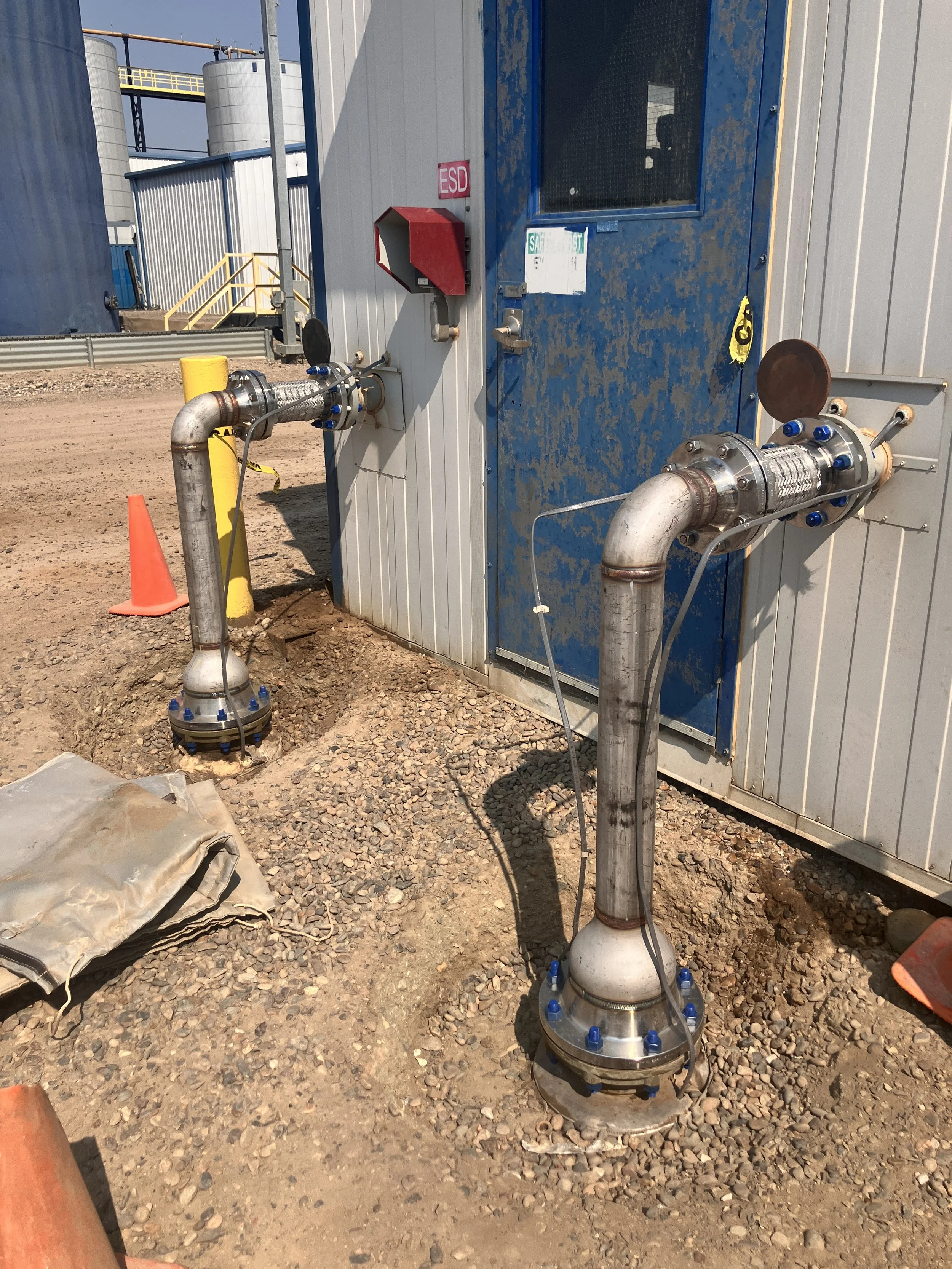 Industrial site with pipe connections and equipment outside, including a pipe with a flange, sensors, and a cabinet with a red caixa, on a gravel surface.