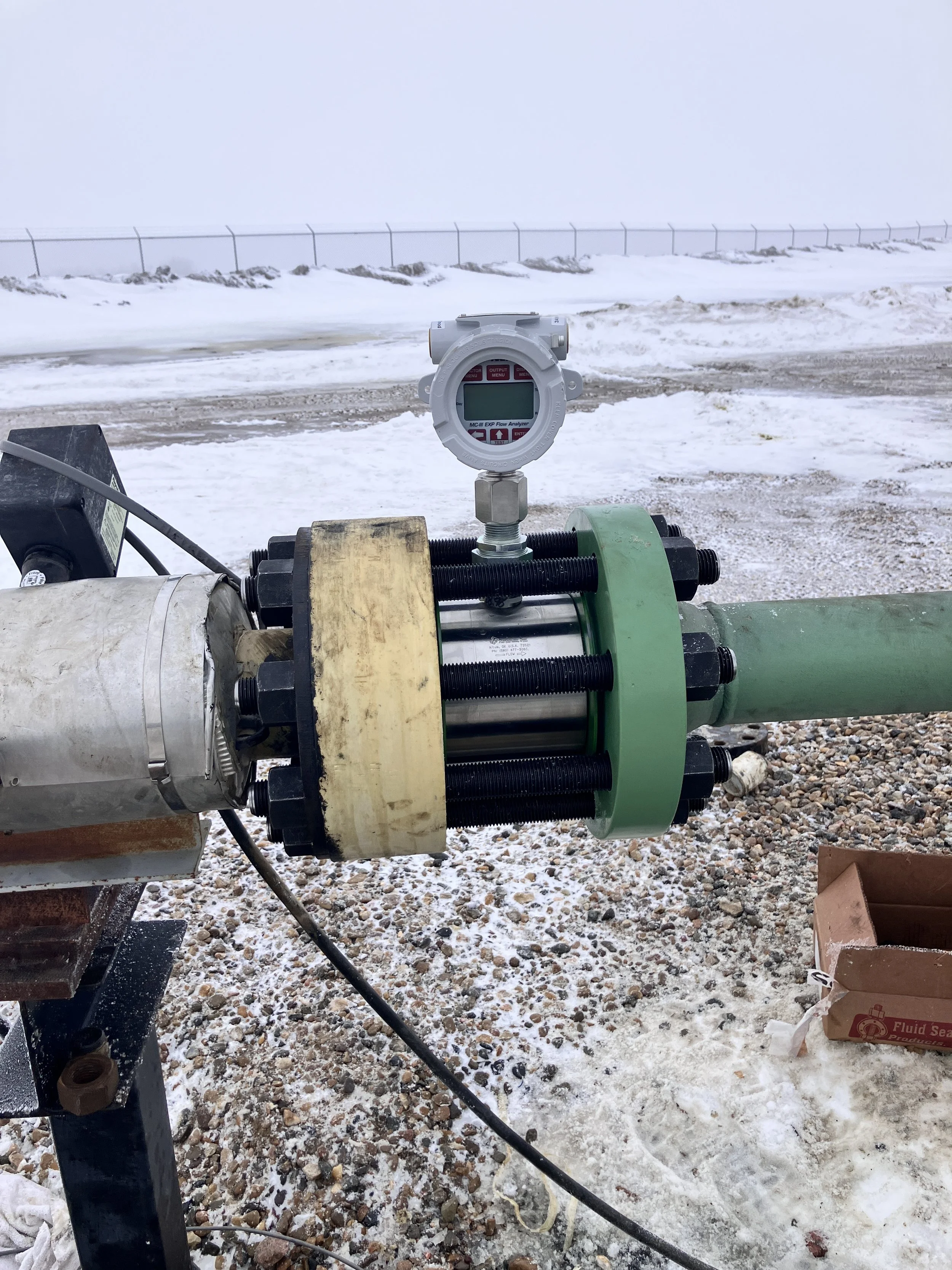 Close-up of a flow analyzer device mounted on a pipeline outdoors, with snow-covered ground and a fence in the background.