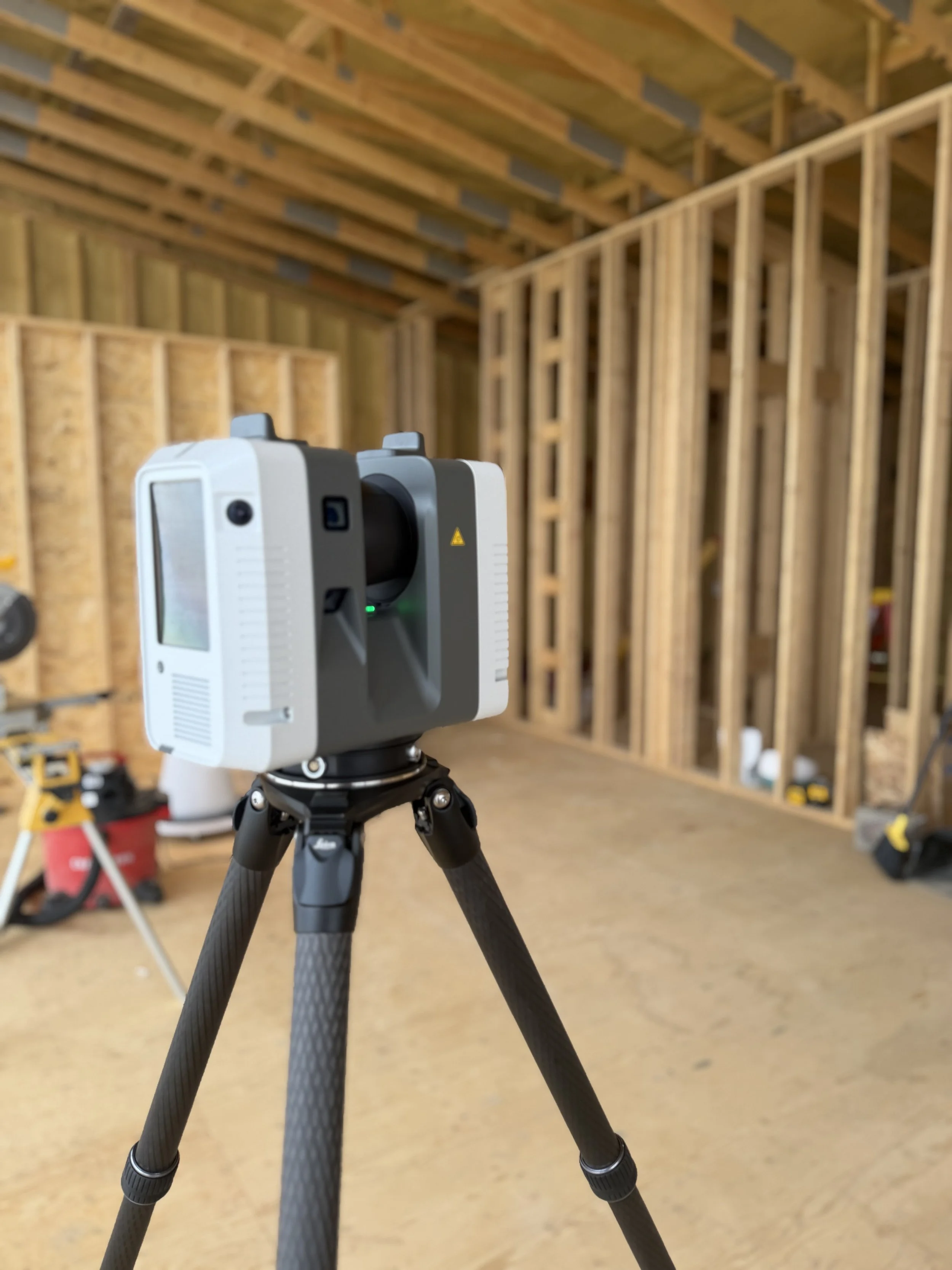 A laser scanner on a tripod inside a residential construction site with unfinished wooden framing for walls and ceiling.