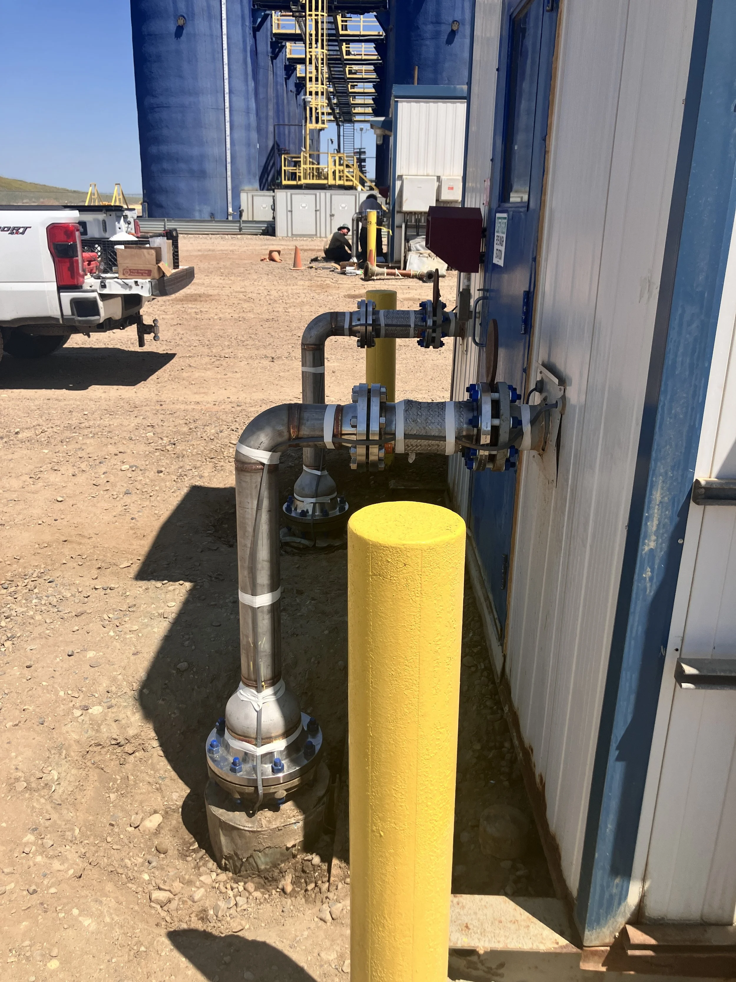 Oil or gas pipelines connected to a white and blue modular containment unit at an industrial site, with a yellow safety barrier and a worker nearby.