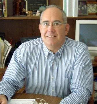 A man with glasses in a striped dress shirt sitting at a desk in an office with books, framed photos, and a computer in the background.