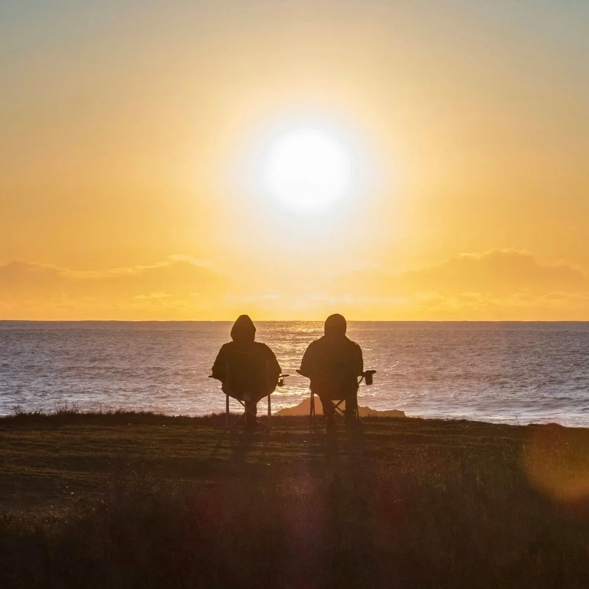 Two people sitting on chairs by the ocean at sunset, gazing at the bright sun over the water with a clear sky.