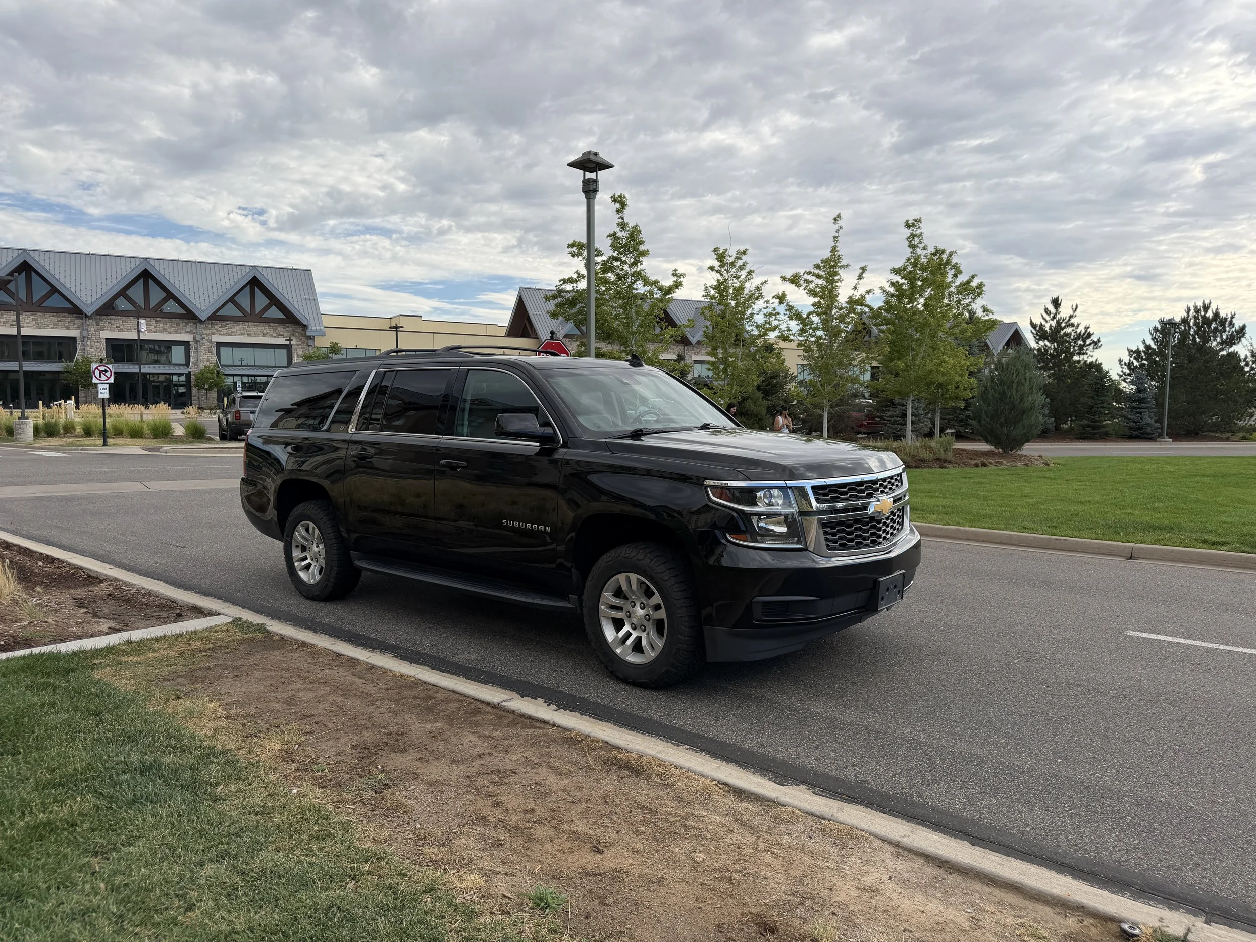 A black Chevrolet Suburban SUV parked in a parking lot with greenery and small trees, a building with a modern roof design, and a cloudy sky in the background.