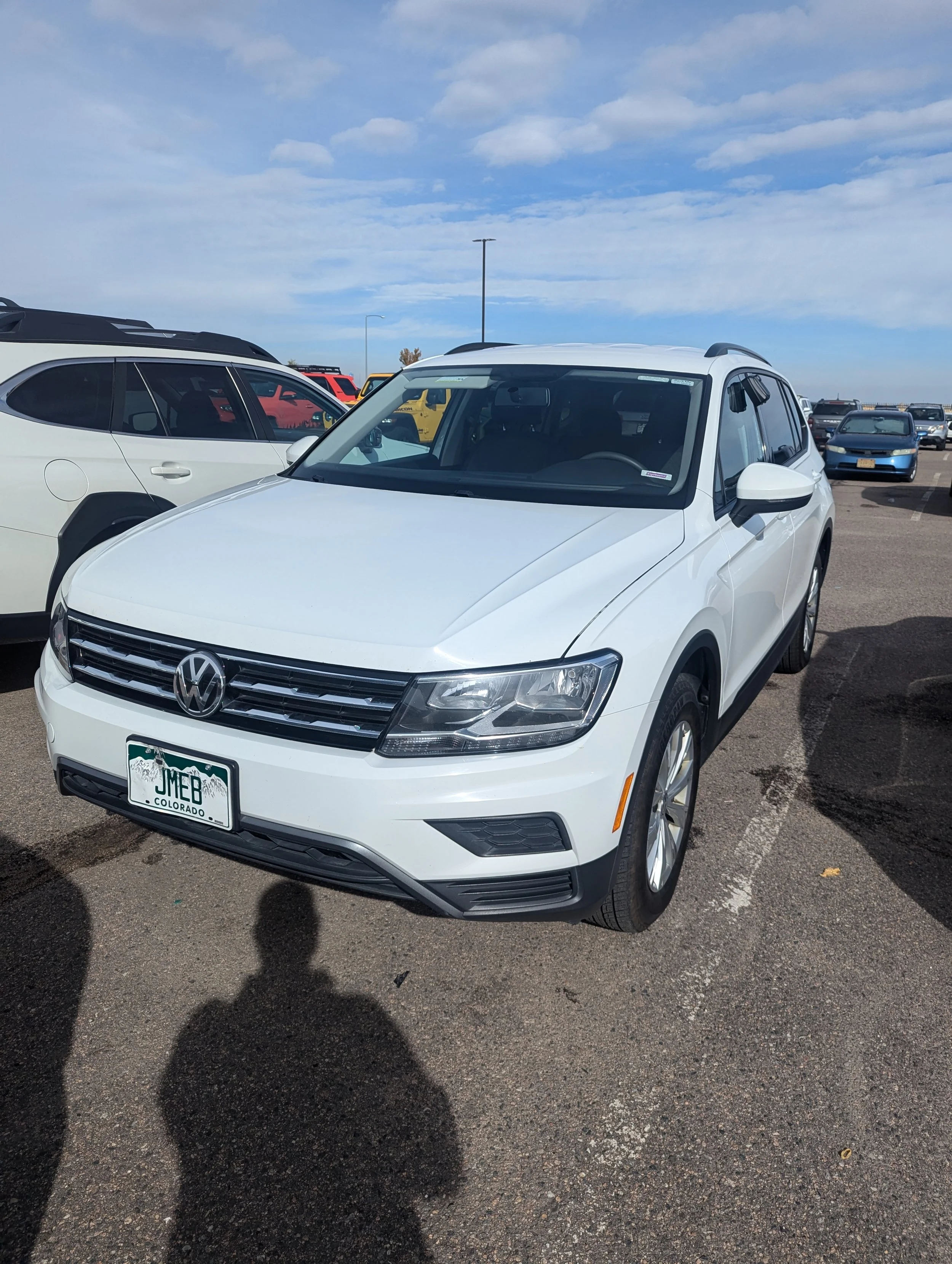 A white Volkswagen SUV parked in a parking lot, with other vehicles around, under a partly cloudy sky.