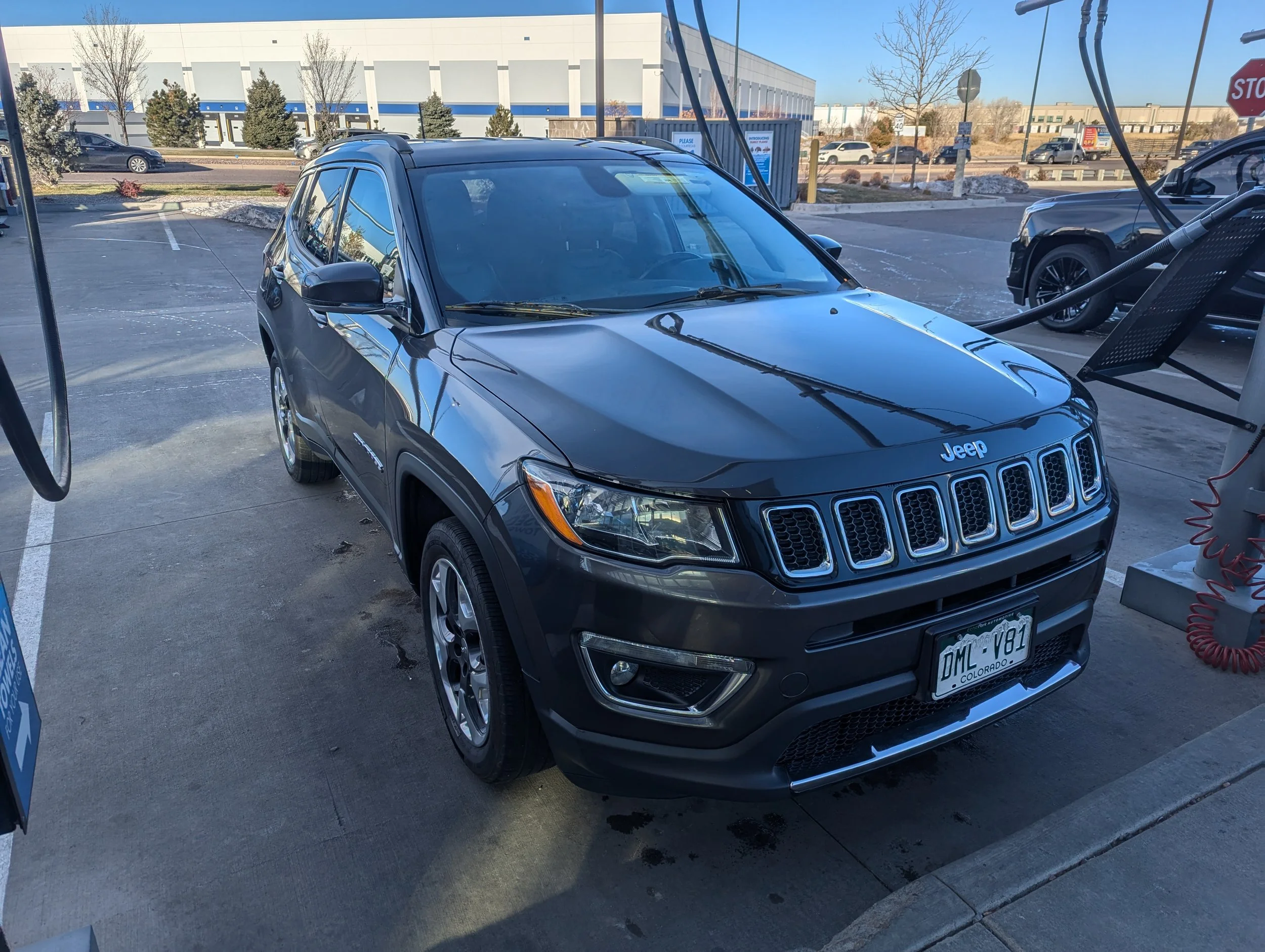 Electric vehicle charging station with a gray Jeep SUV plugged in, set in a parking lot with a large building and some trees in the background.