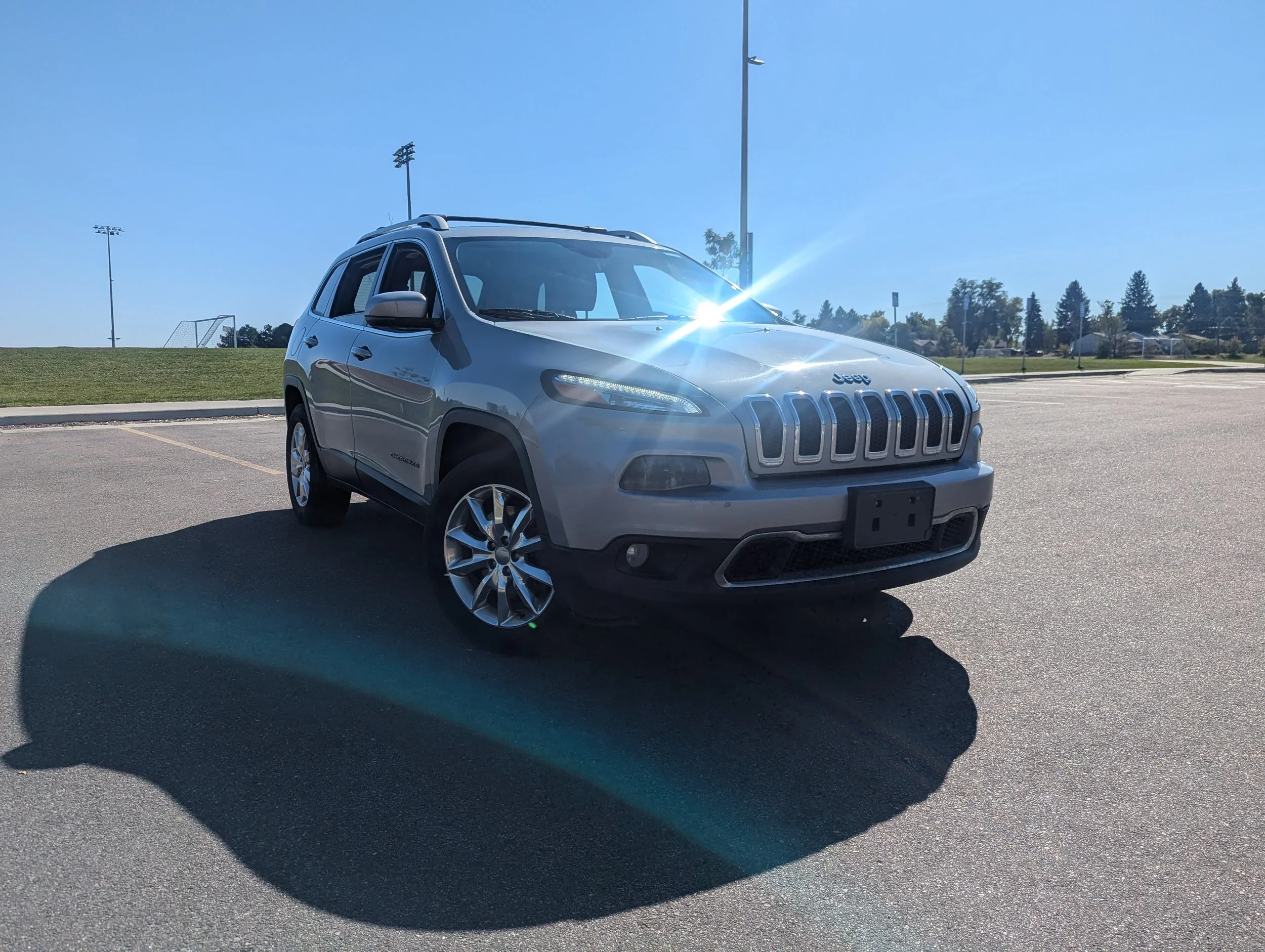 A silver Jeep SUV parked in an empty parking lot on a sunny day, with football fields and trees in the background.
