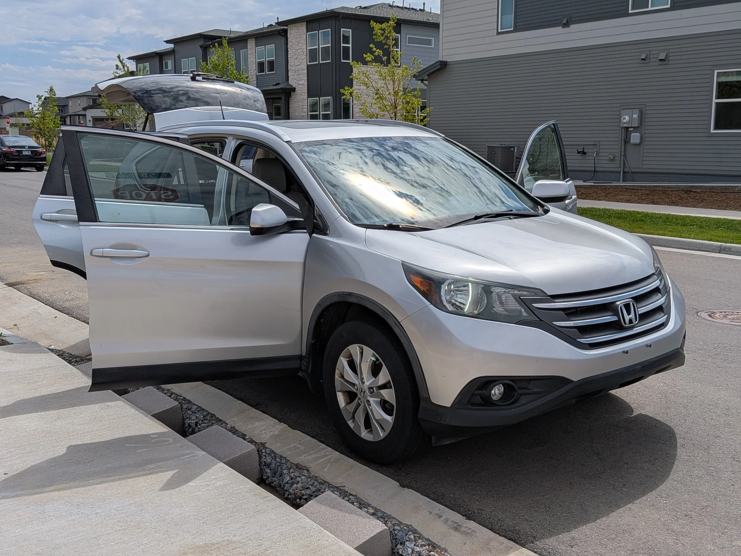 A silver Honda CR-V with open front door and open rear hatch parked on a residential street with townhouses in the background.