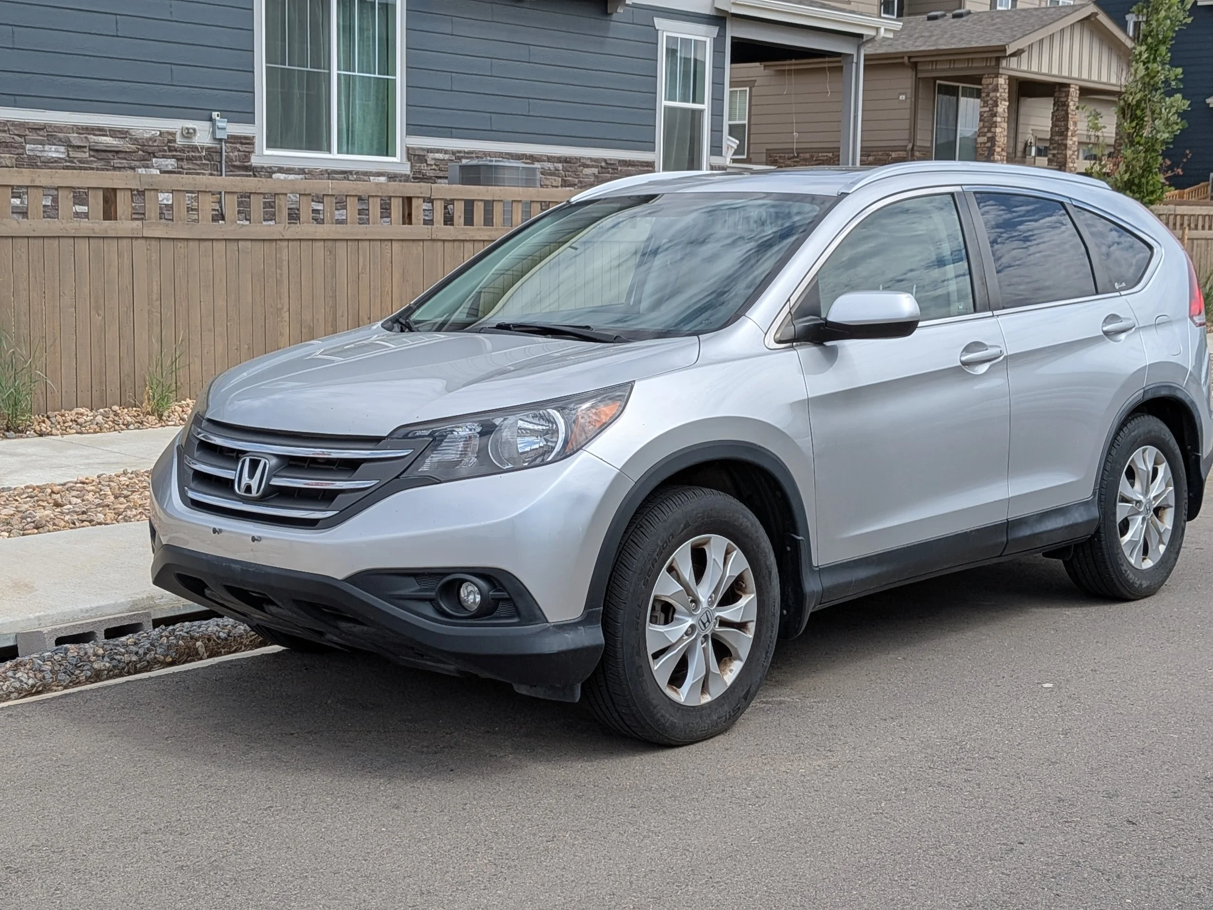 A silver Honda SUV parked on a residential street in front of a house with blue siding and stone accents, and a wooden fence.