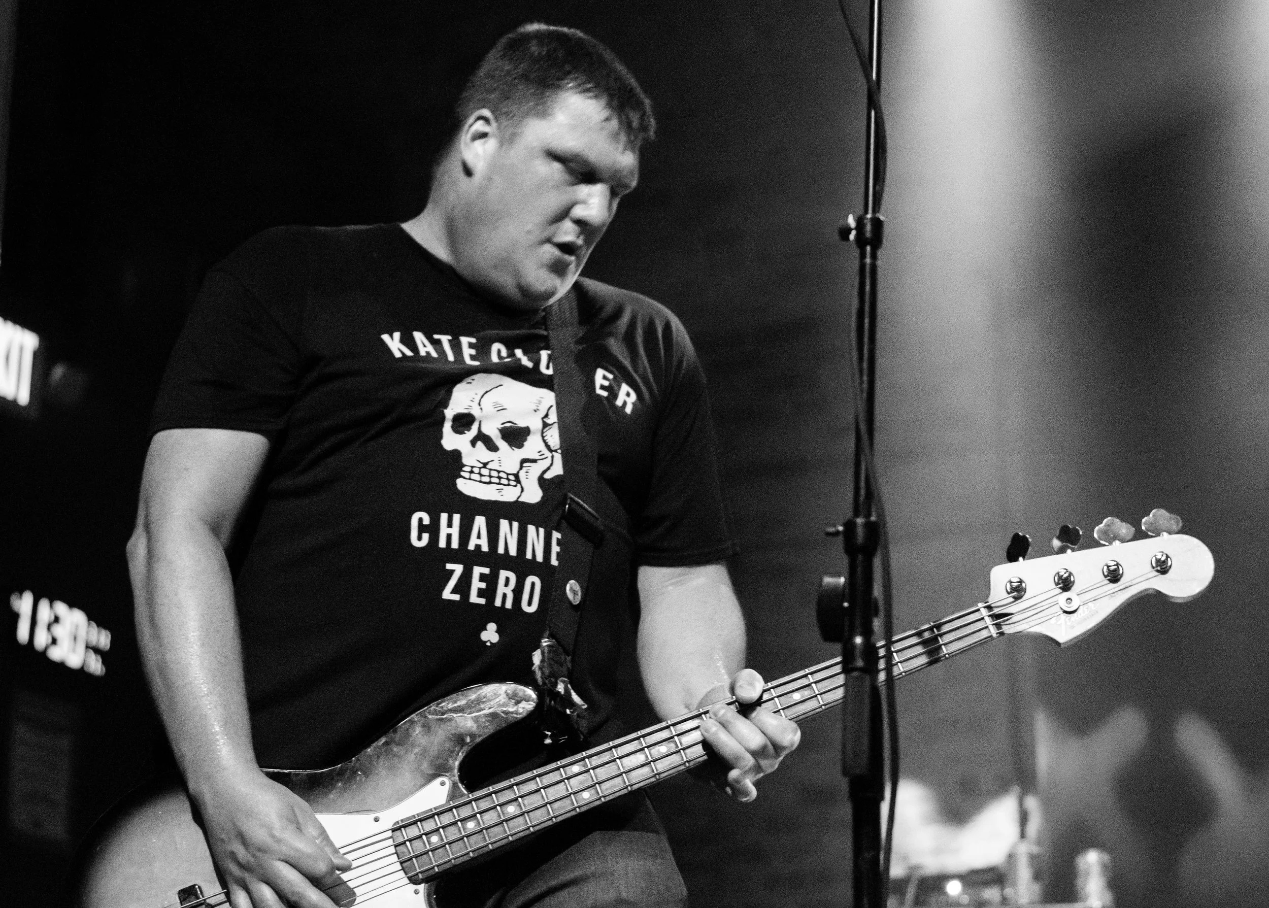 A black and white photo of a young person playing an electric bass guitar, with a T-shirt that has a skull graphic and text reading "KATE GORZER CHANNEL ZERO". The person appears to be performing or practicing, with a music stand nearby and a microph
