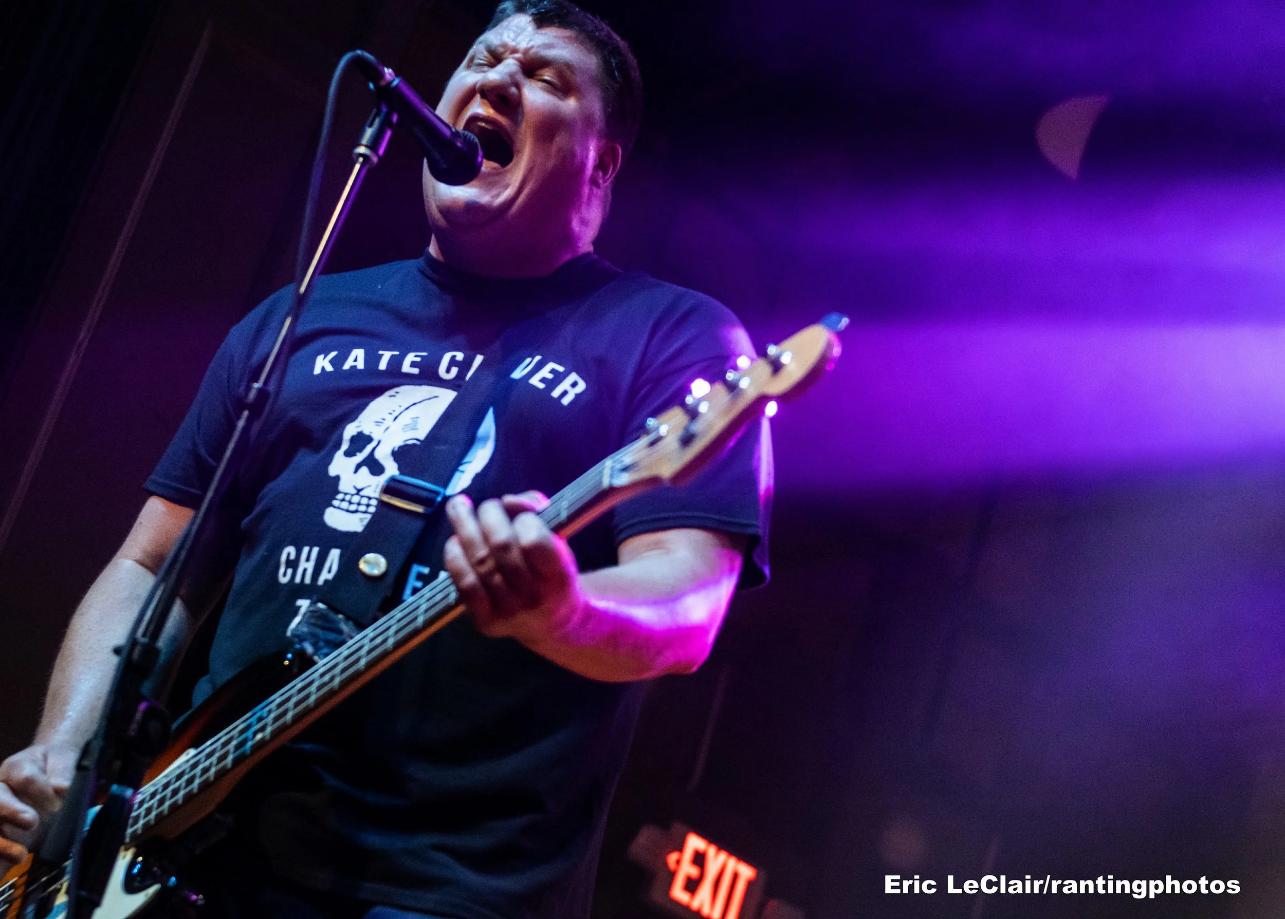 A person passionately singing into a microphone while playing an electric guitar with purple lighting in the background.