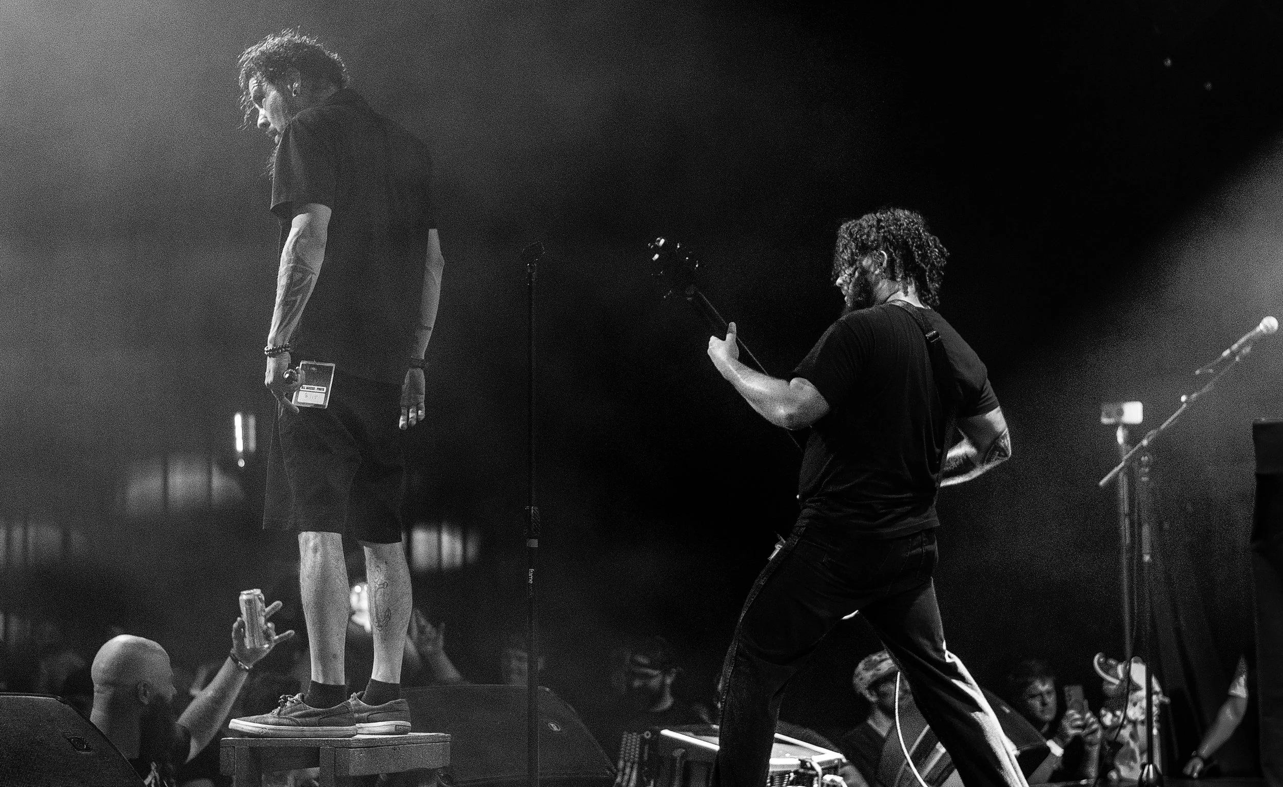 A black and white photo of a musician on stage with another person who is holding a microphone and a guitar. The musician is standing on a platform, looking down, while the other person appears to be playing the guitar. The stage has microphones and 