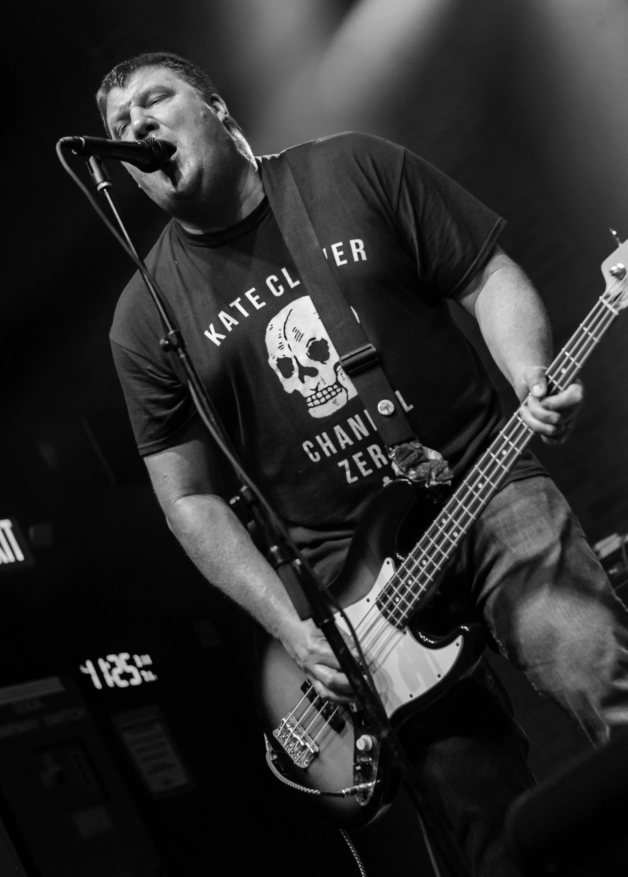 A black and white photo of a man singing into a microphone while playing an electric bass guitar. He is wearing a T-shirt with a skull graphic and the words 'KATECH' and 'CHANN' are visible on the shirt.