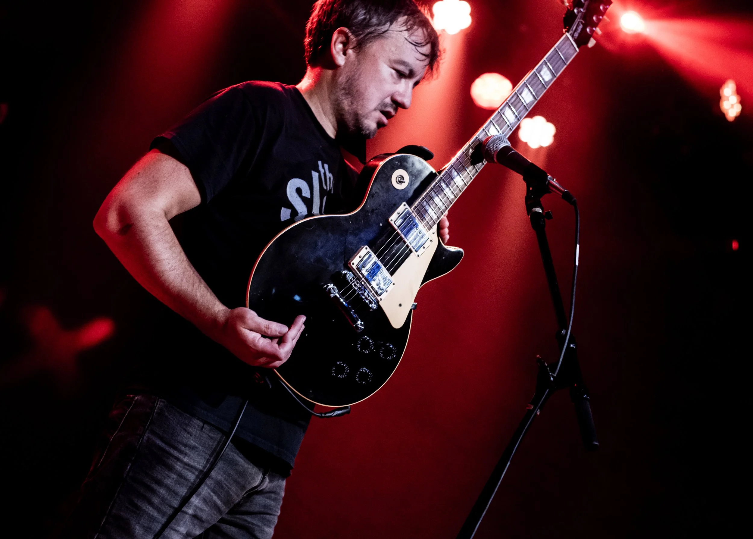 A man with short hair and a beard playing an electric guitar on stage, surrounded by red stage lights.