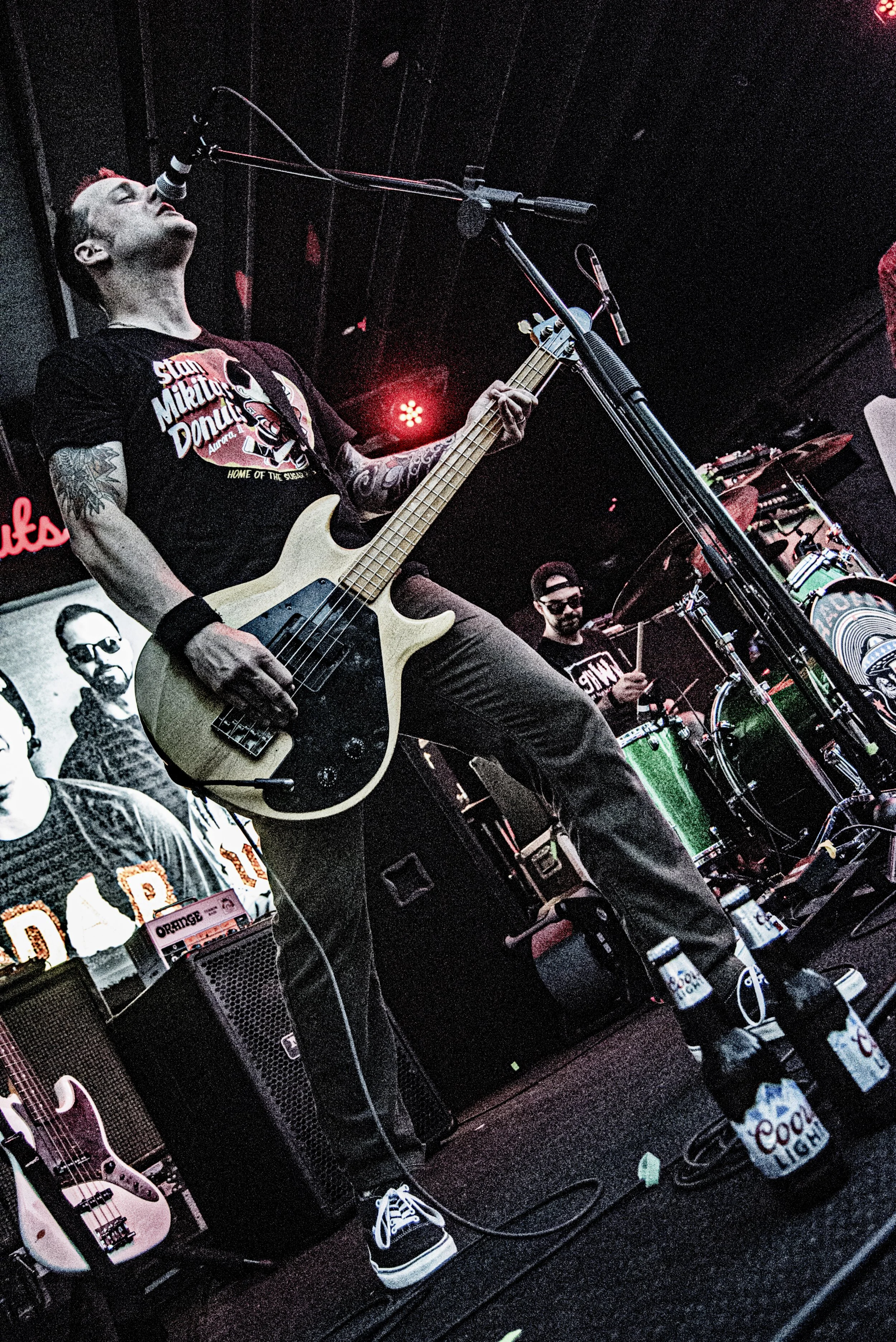 A musician with tattoos on his arm, wearing a black T-shirt with a logo, playing a bass guitar on stage. There is a microphone in front of him, and a drummer is visible in the background. The stage is dimly lit with red lights.