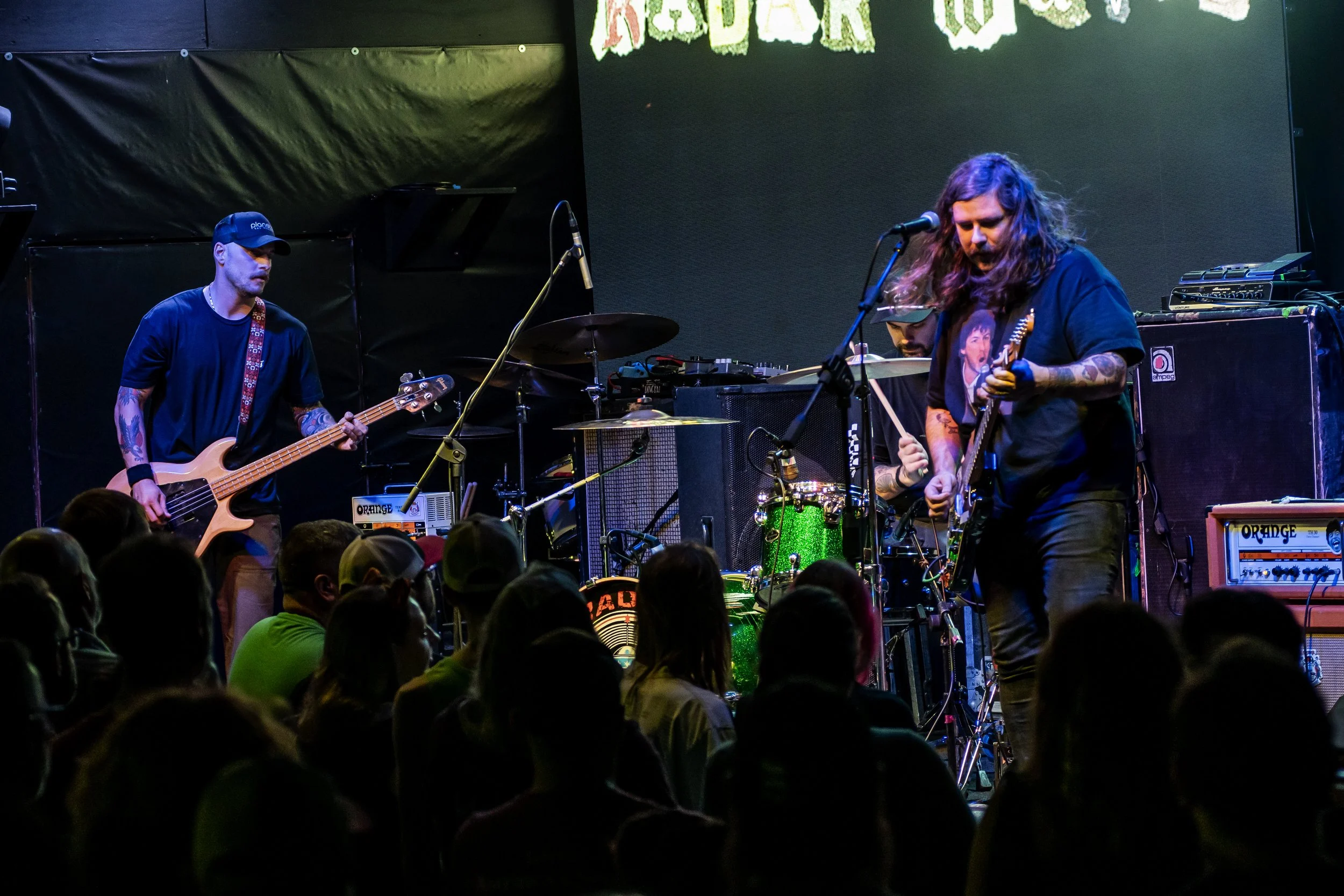 A band performs on stage with three members playing instruments. The crowd is visible in the foreground, watching the performance. The stage backdrop features a large screen with partially visible text.
