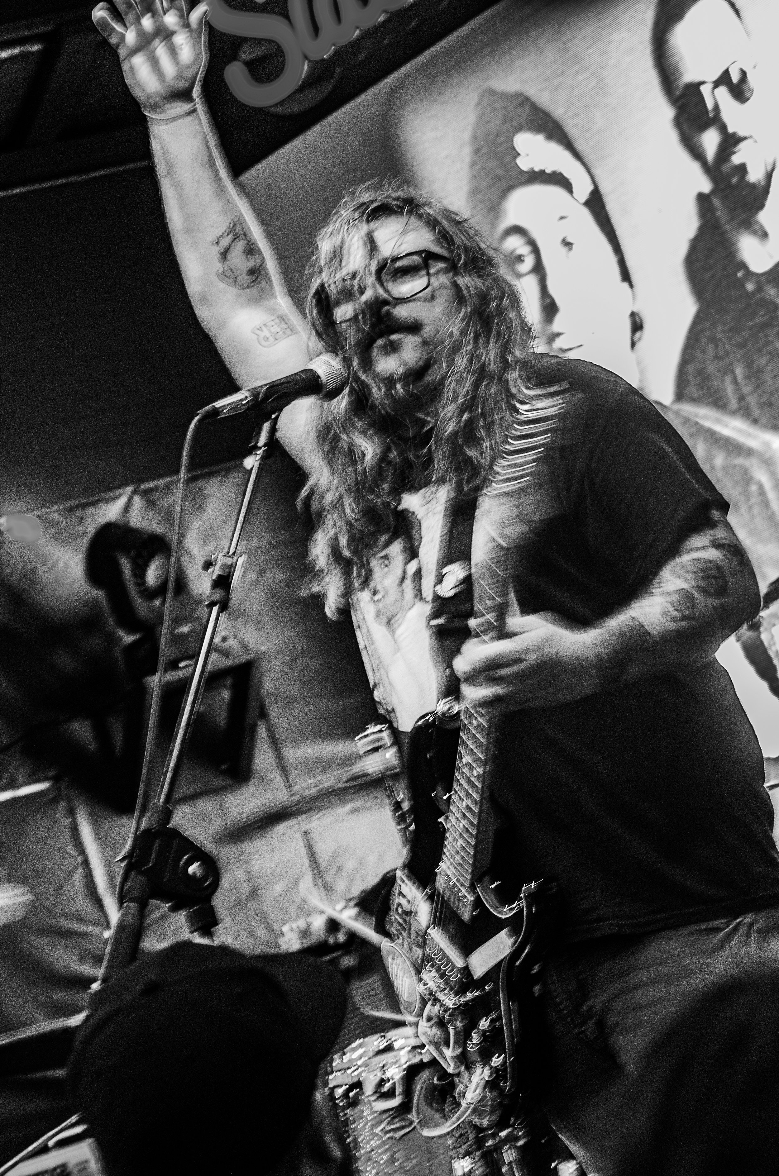 A person with long hair, glasses, and tattoos playing guitar and singing into a microphone in a black and white photo. Behind them, there is a projected image of a person's face on the wall.