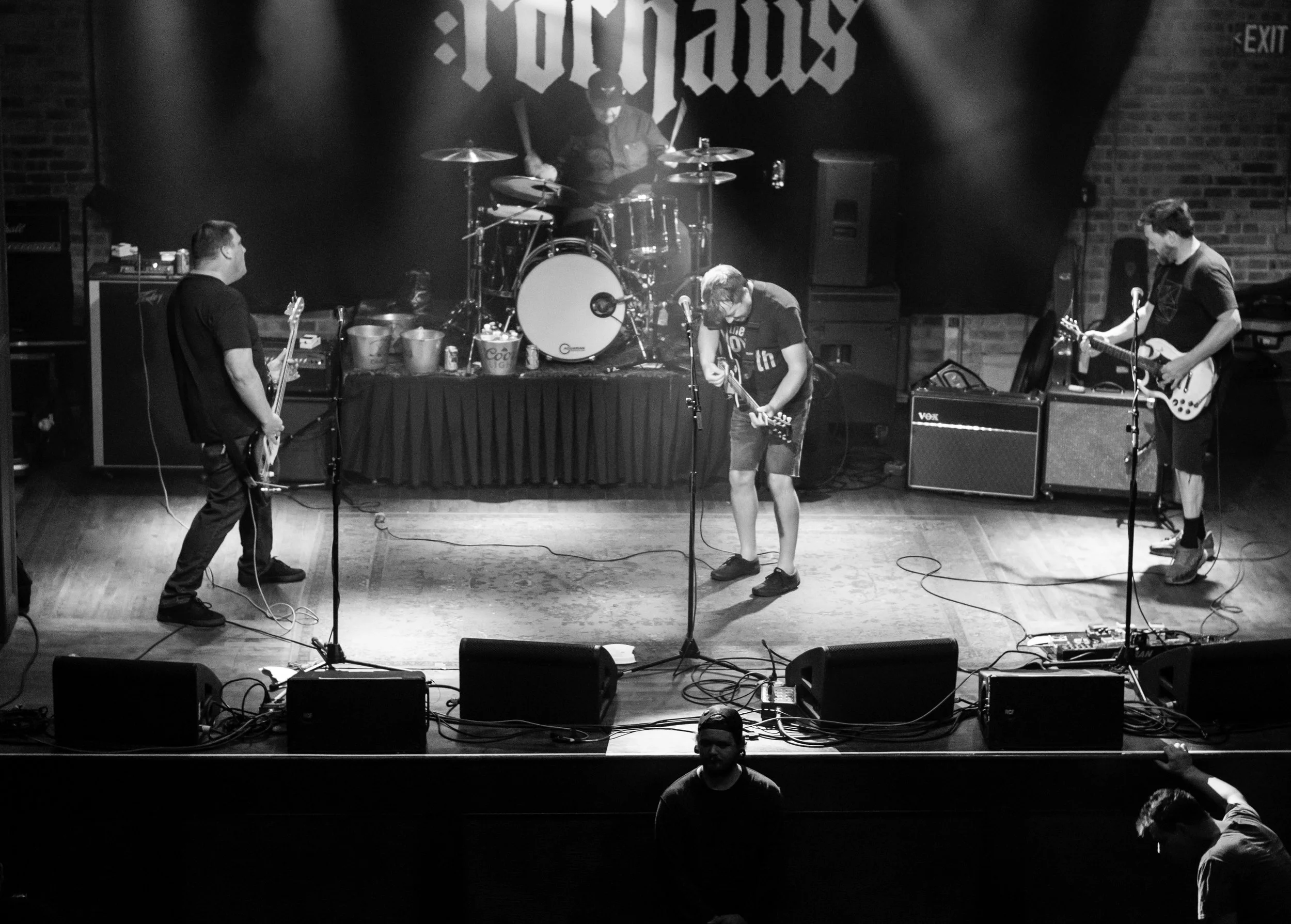 Black and white photo of a band performing on stage, with four members playing instruments and a drummer behind them. There is a large banner with the word 'TOXICUS' hanging on the back wall. Audience members are visible in the foreground.