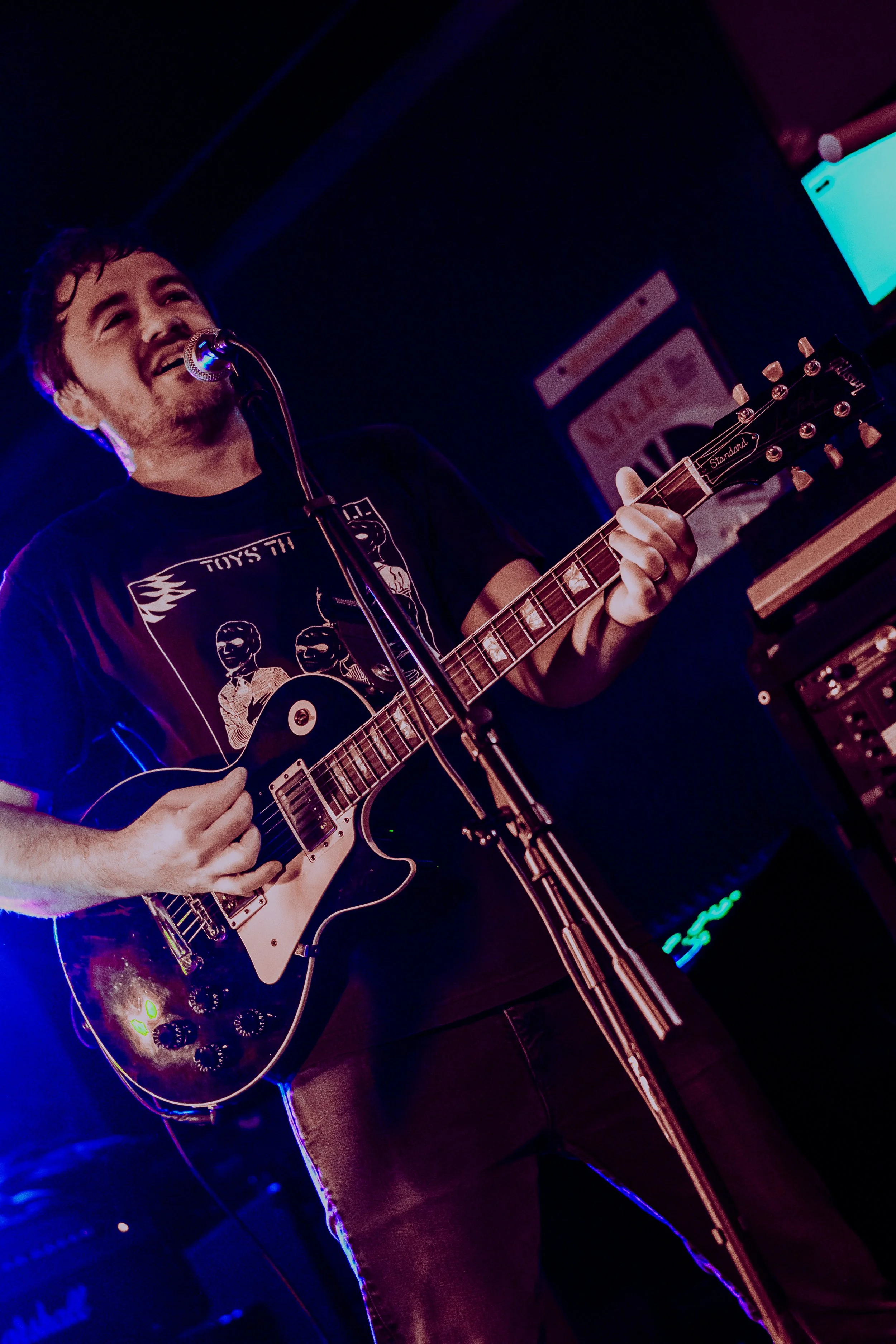 Male musician playing an electric guitar, singing into a microphone in a dimly lit venue with blue and purple lighting.