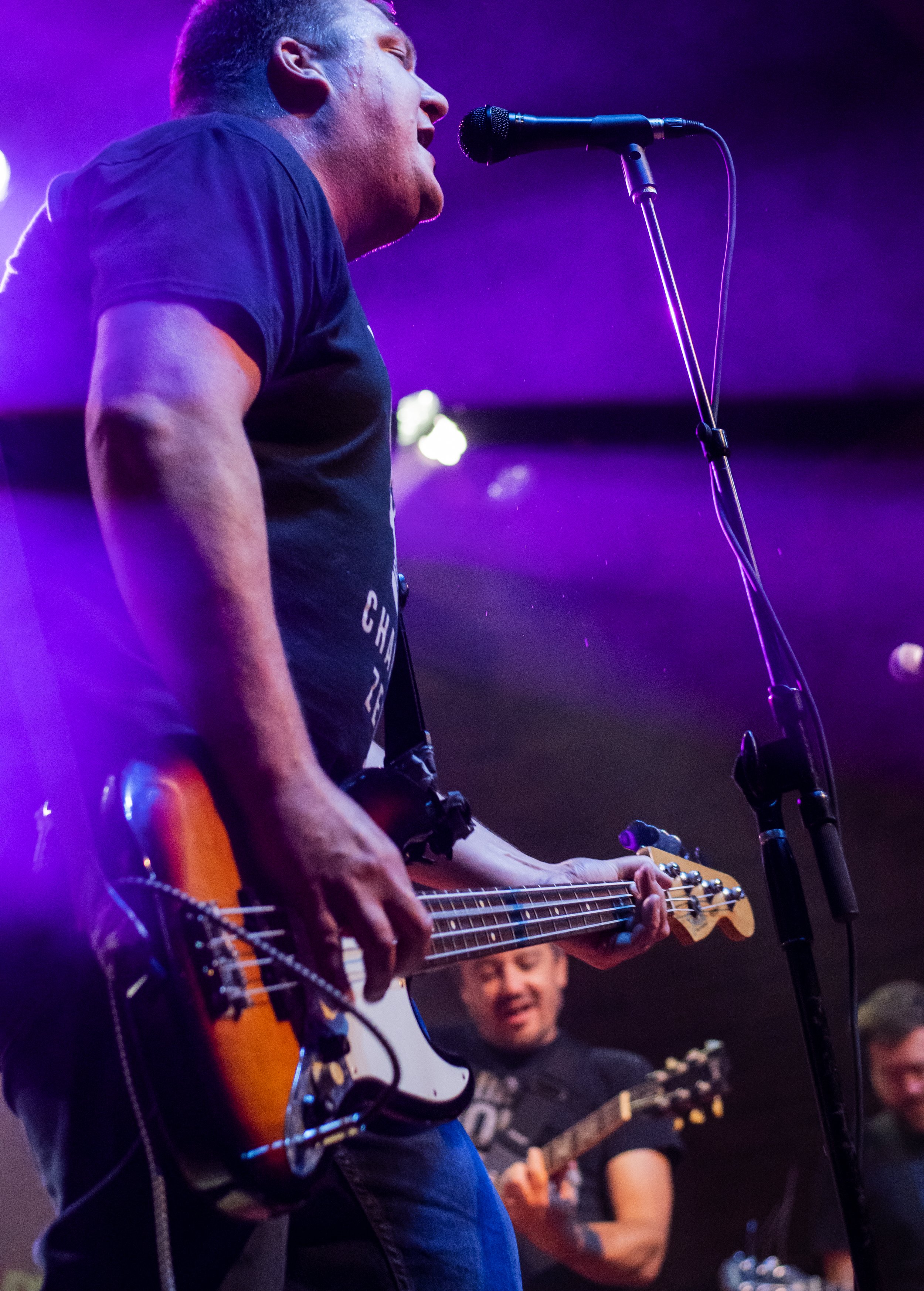 Musician singing into microphone while playing electric guitar on stage with purple lighting, accompanied by other band members.