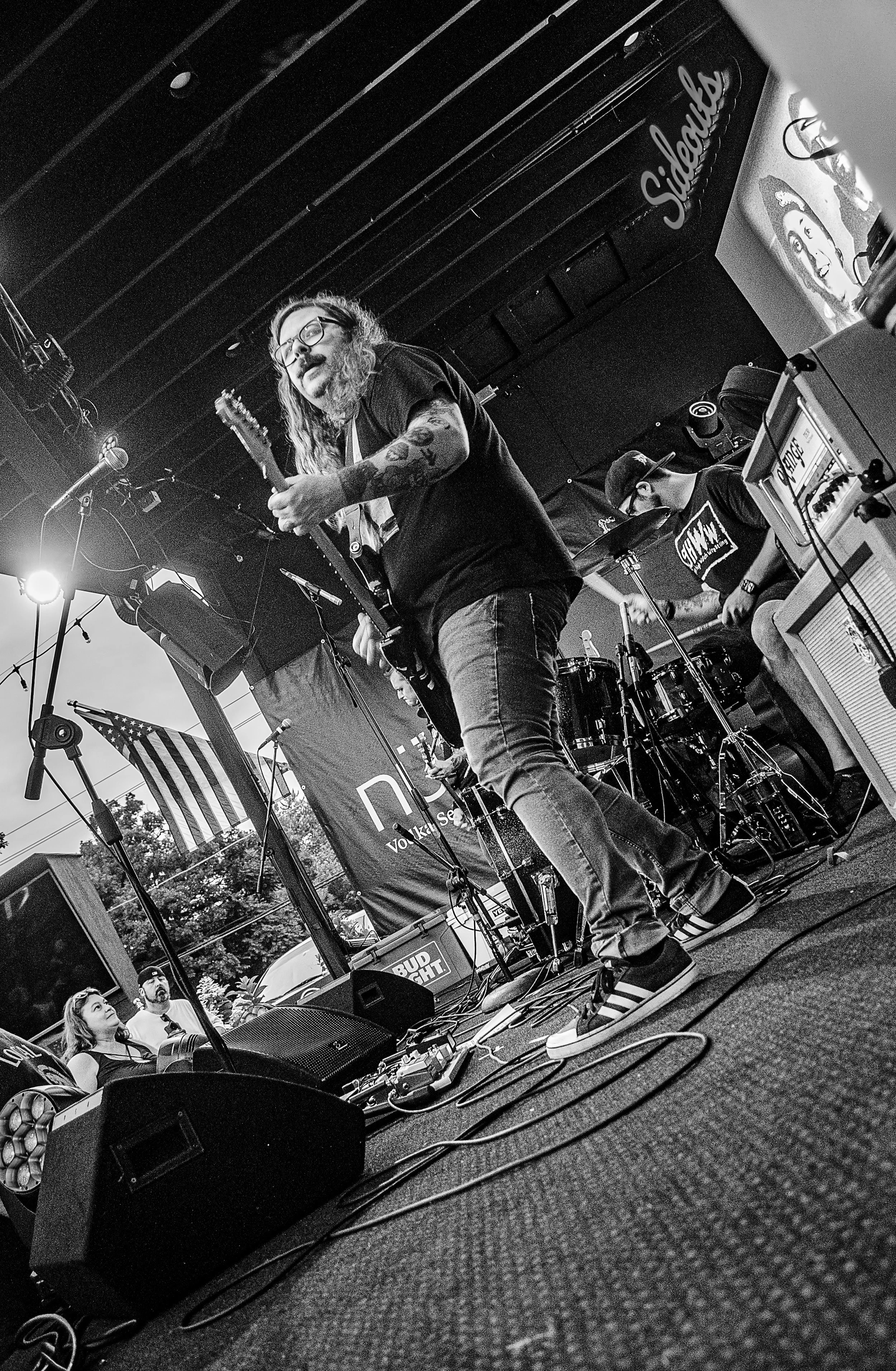A man with glasses and long hair playing an electric guitar on stage during a live performance. There is a drum set behind him, and a woman and a man are watching the performance from the audience. The stage has various equipment, and a sign with the