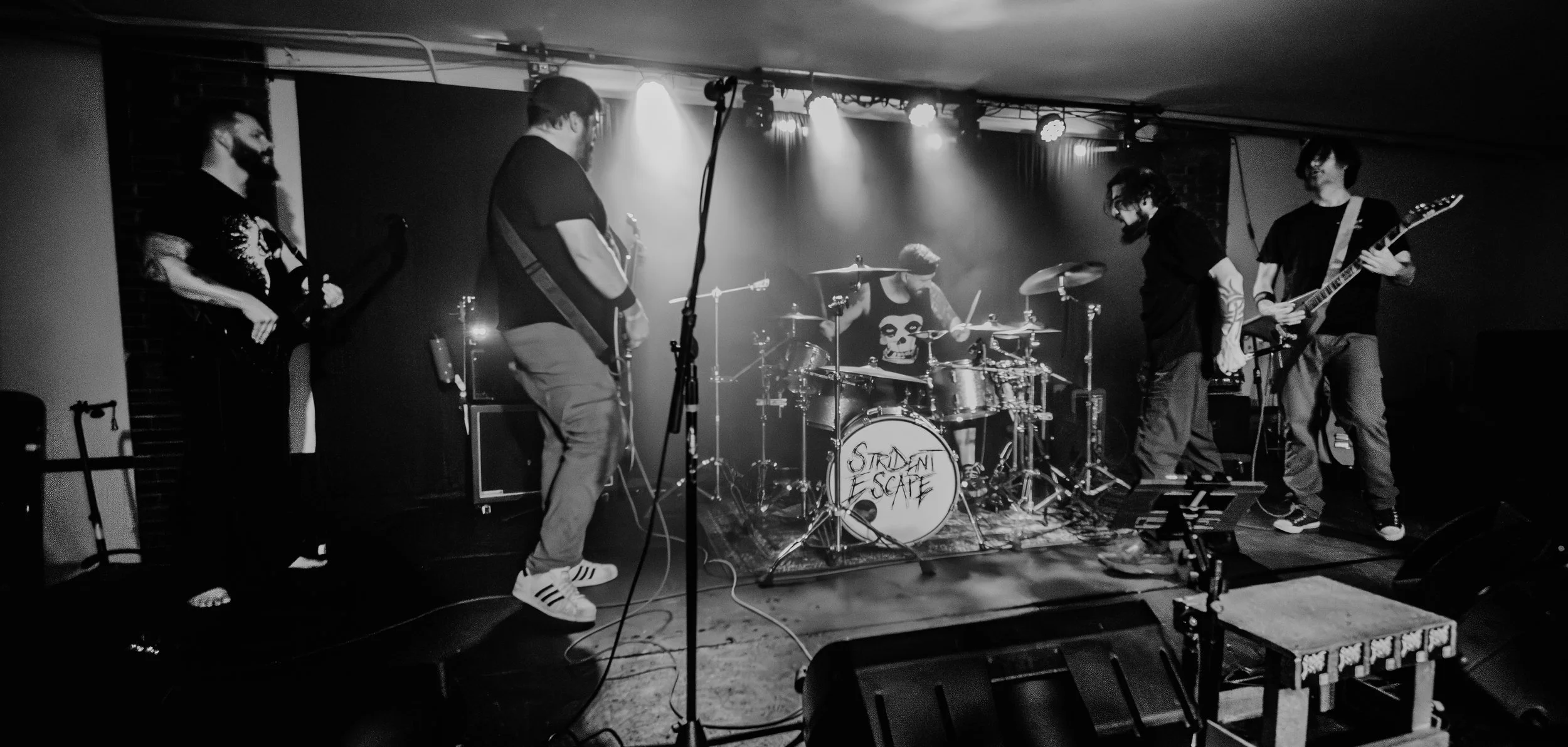 Black and white photo of a band performing live on stage. The band has five members, including a drummer with a skull shirt, guitarists singing, and others playing guitars. The drum set has the words "Stray Escape" written on the bass drum. Stage lig