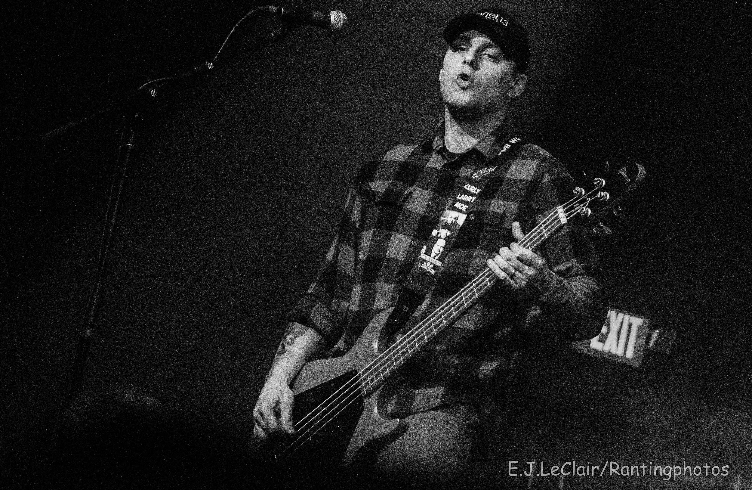 A person playing an electric guitar and singing into a microphone on a stage. The person is wearing a plaid shirt, a cap, and has a lanyard around their neck with various patches. The background is dark with an exit sign visible.