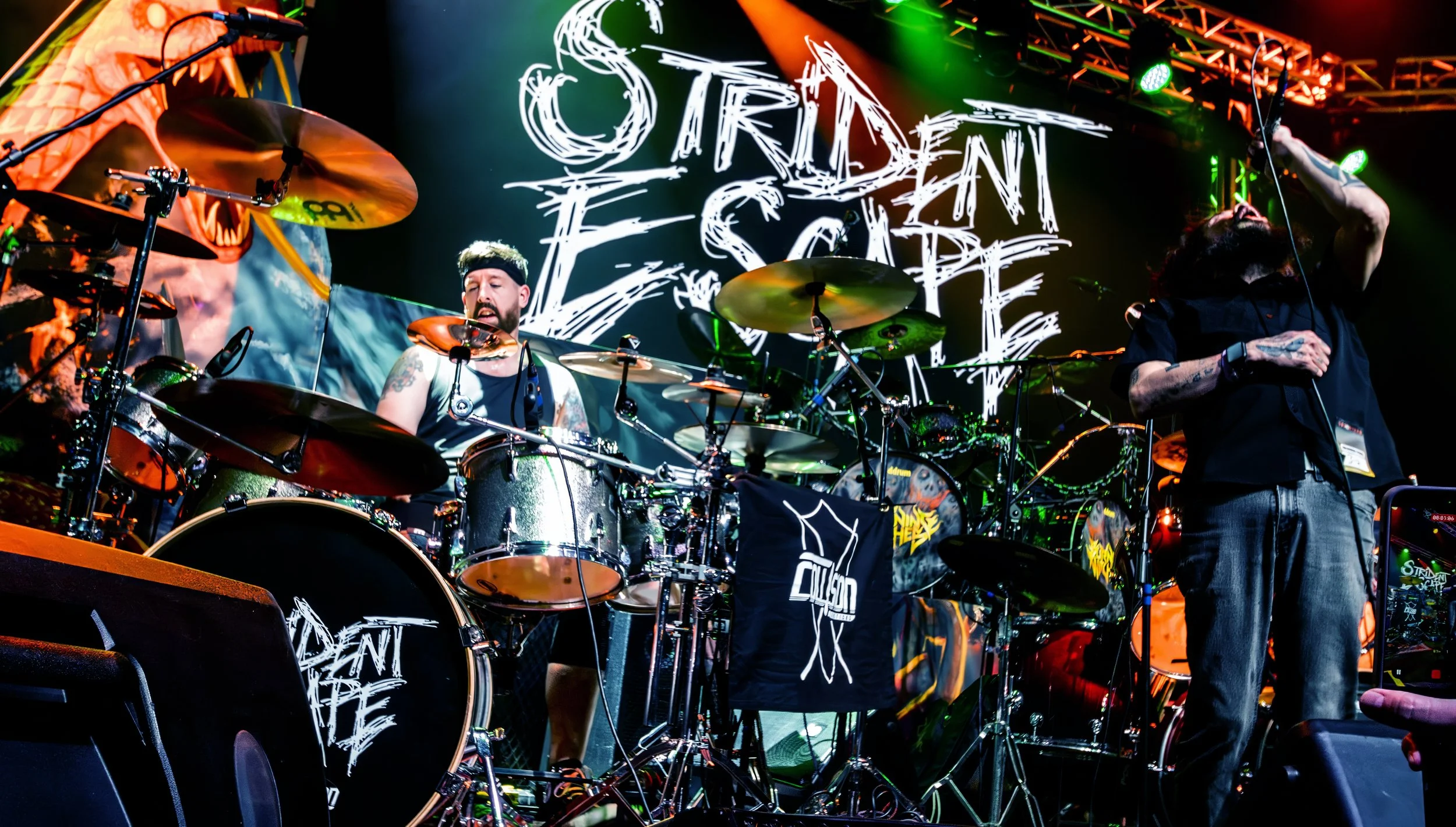 Musicians performing on stage during a concert with a large screen displaying 'Orion Fest' in the background. The drummer is visible with a drum set, and another musician is mid-performance.