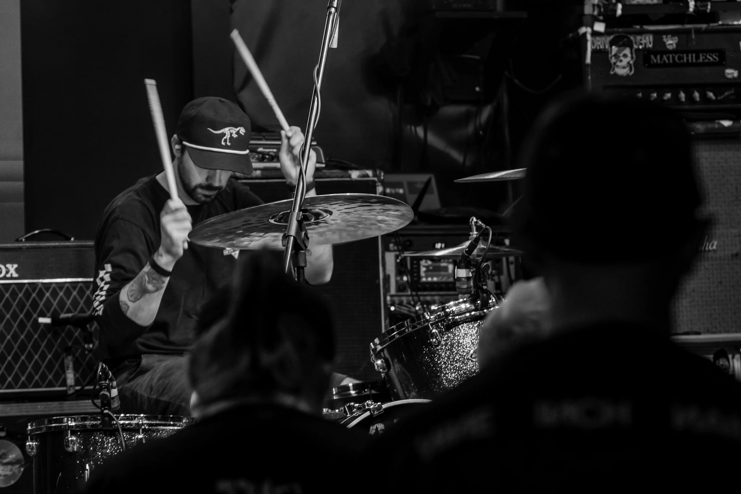 A man playing drums in a music studio, wearing a hat with a dinosaur logo and tattoos on his arm, with music equipment and speakers in the background.