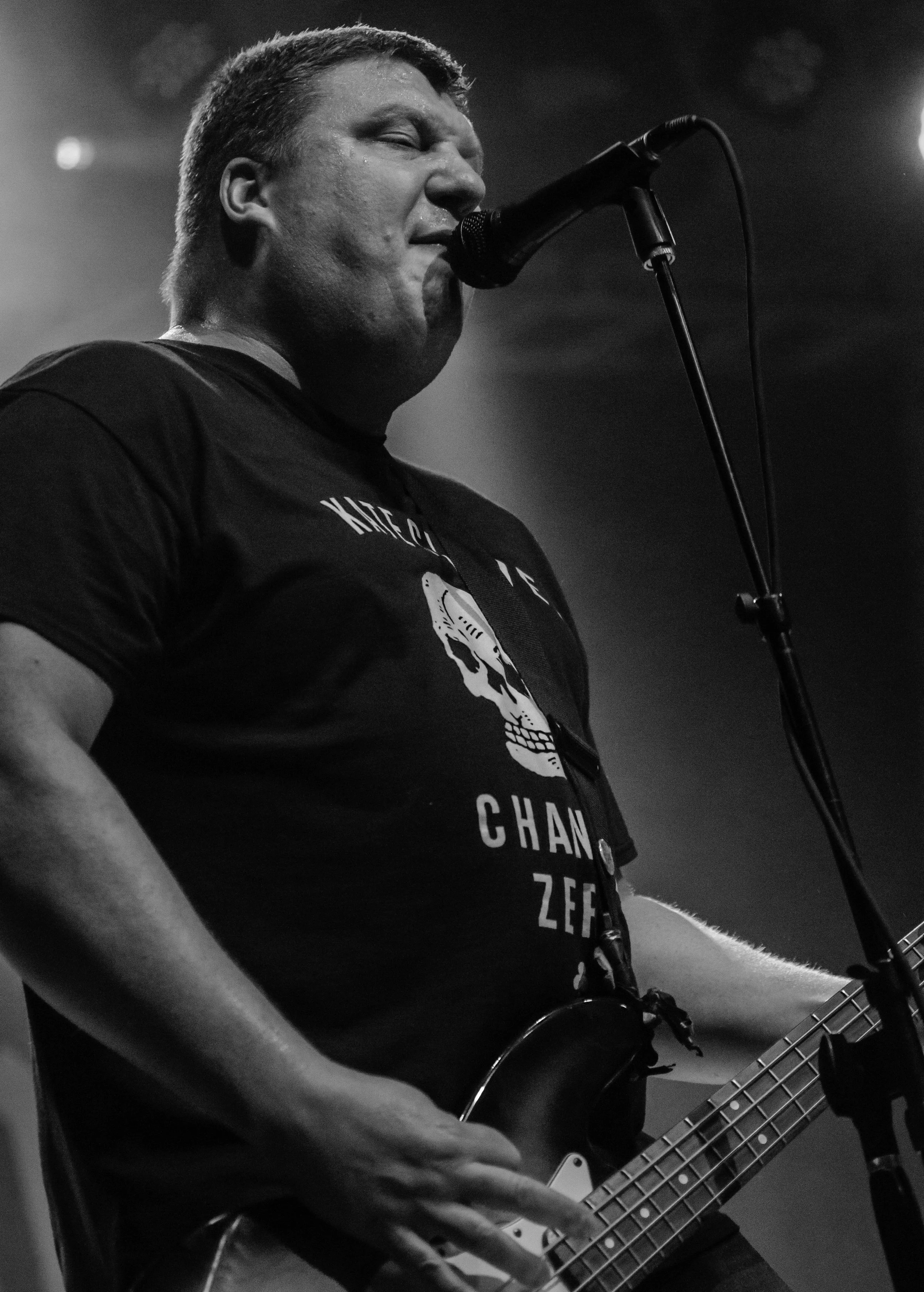 A black and white photo of a man singing into a microphone while playing an electric guitar. He is wearing a T-shirt with a skull graphic. The man has short hair and appears to be performing on stage.