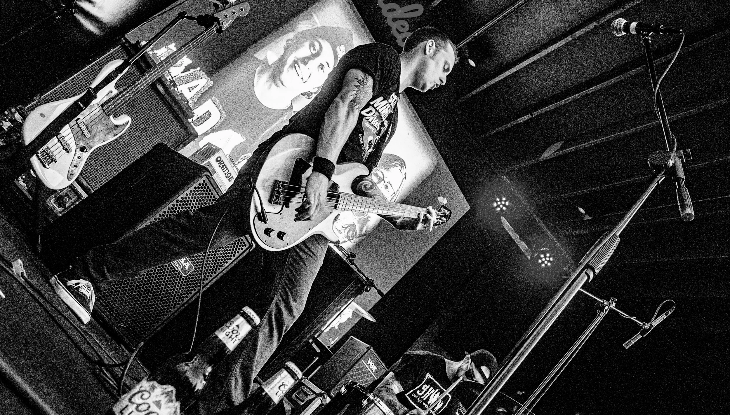 A musician playing an electric bass guitar on stage with a microphone stand nearby. The stage has various guitar amplifiers and pedals, and a banner with artwork and text in the background.