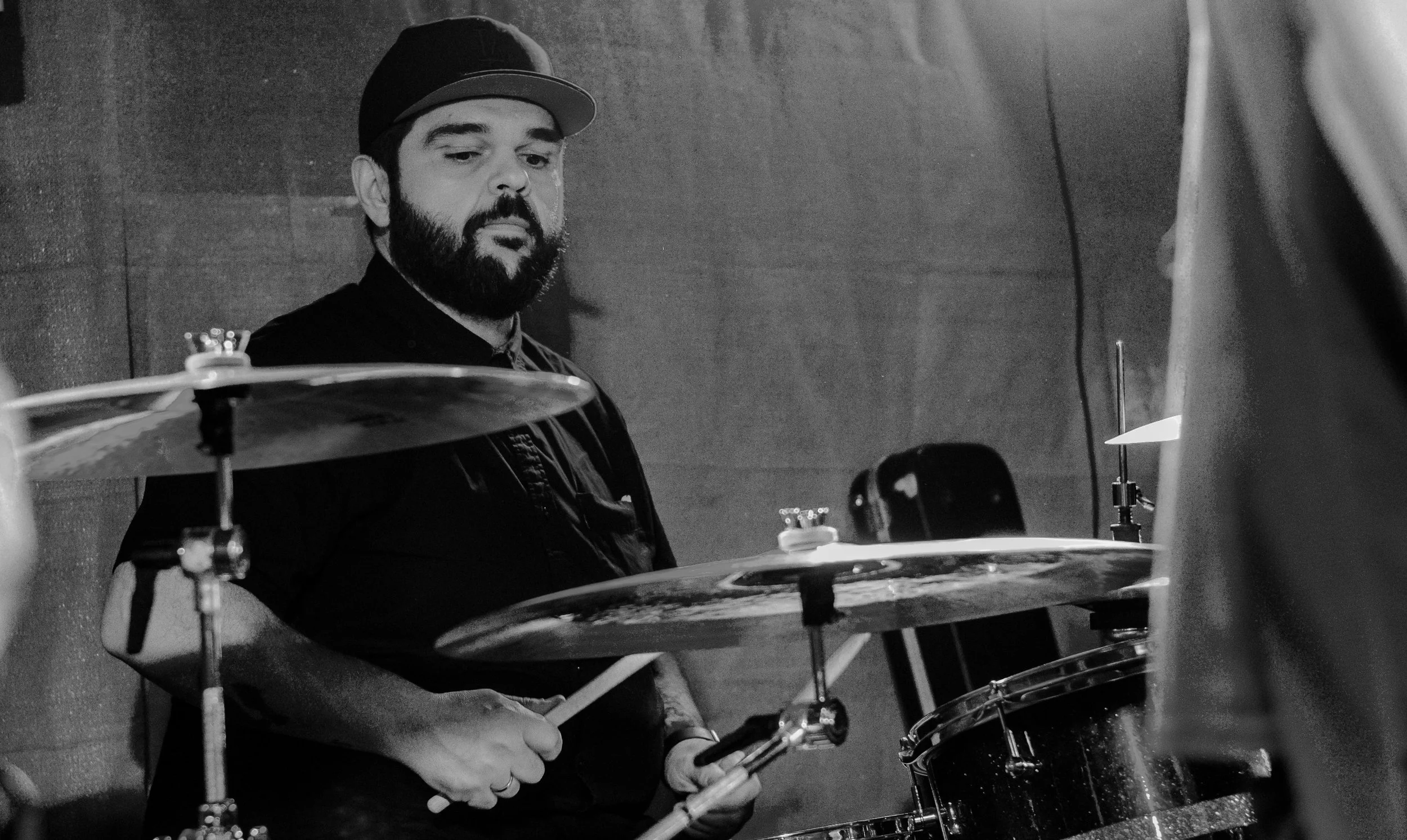 A man with a beard wearing a cap playing the drums in a studio, captured in black and white.