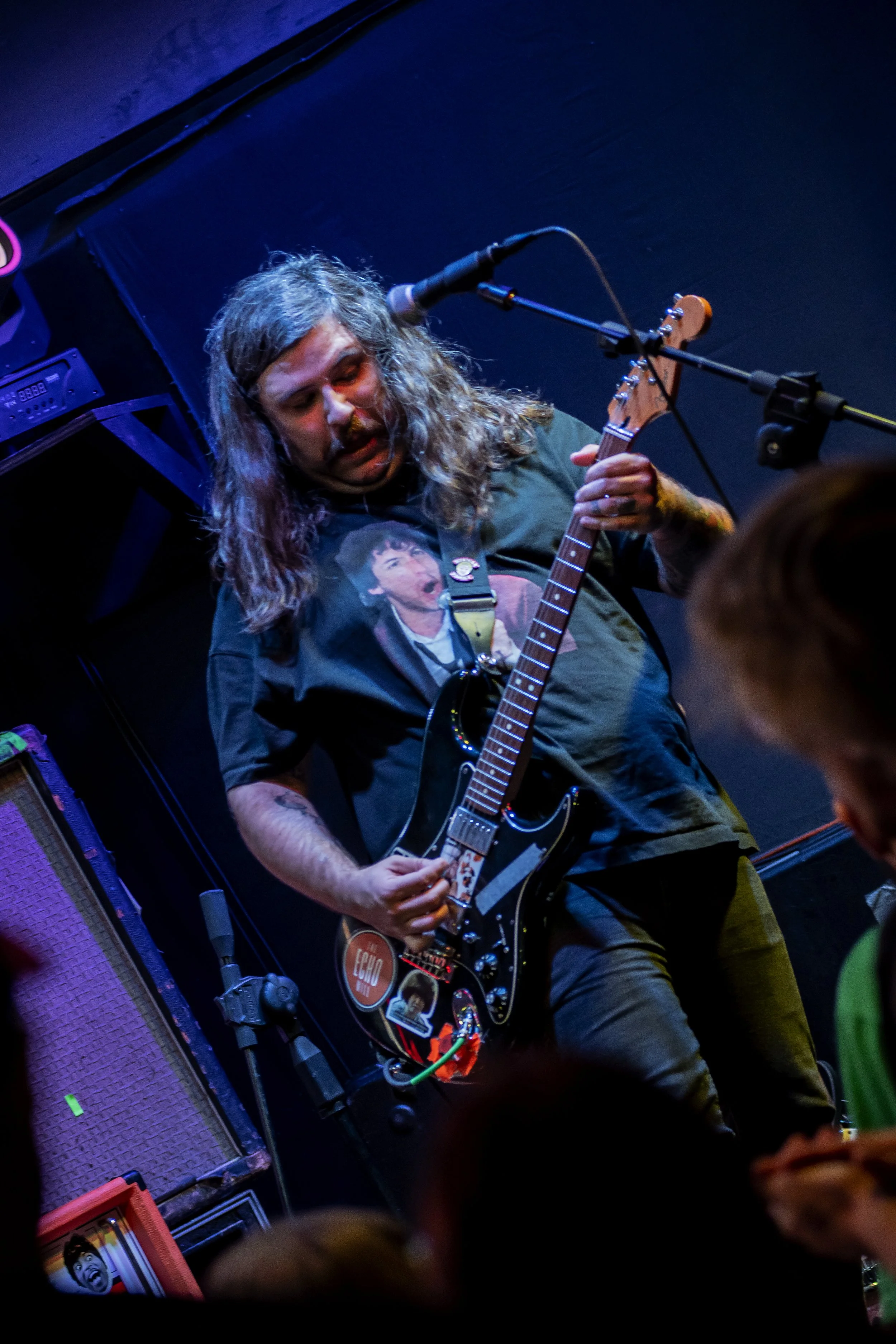 A male musician playing an electric guitar on stage, wearing a black T-shirt with a graphic print of a person's face, and surrounded by musical equipment and dark blue background.
