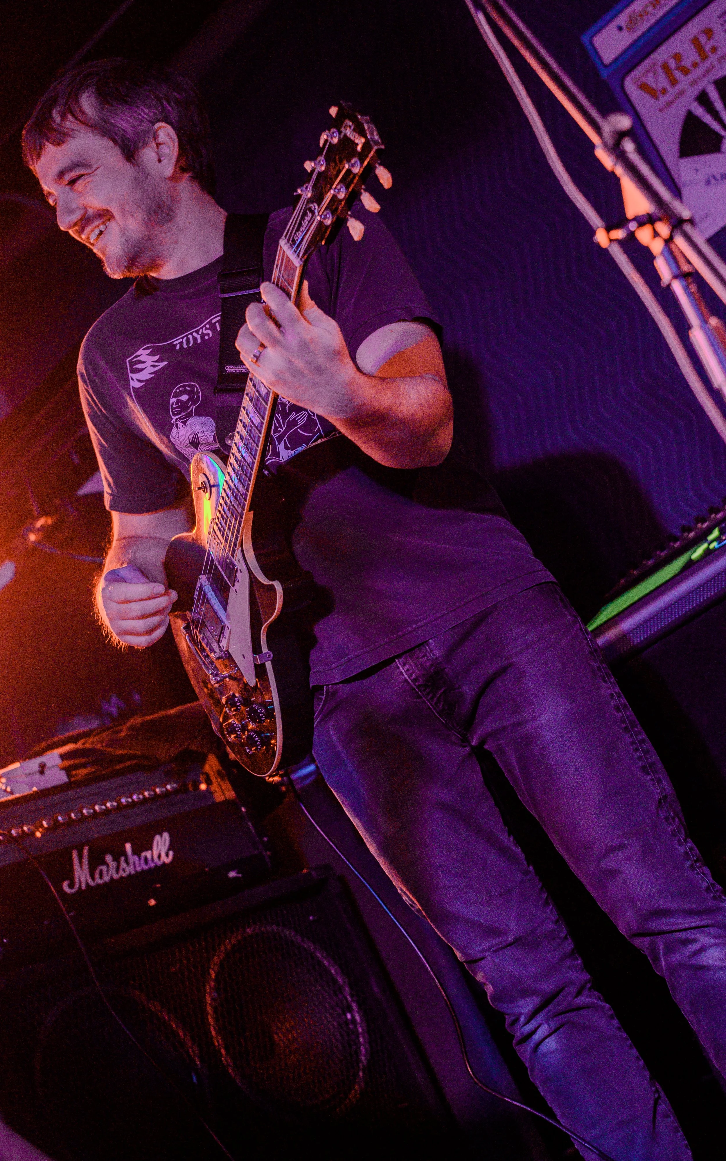 A man playing an electric guitar, smiling, with Marshall speaker and other musical equipment in a dimly lit setting.
