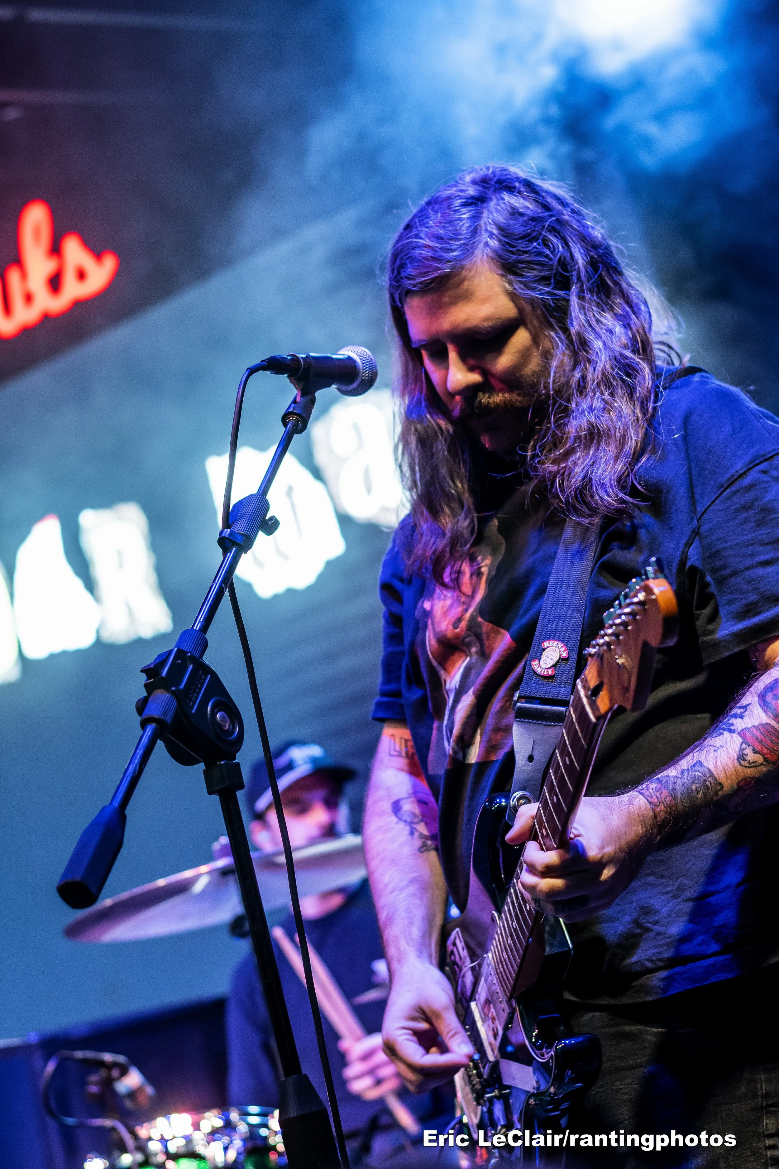 A musician with long hair and tattoos playing an electric guitar on stage, with a microphone in front of him, and a band member playing drums in the background. Stage lights are visible, and there is neon signage partially visible on the wall.