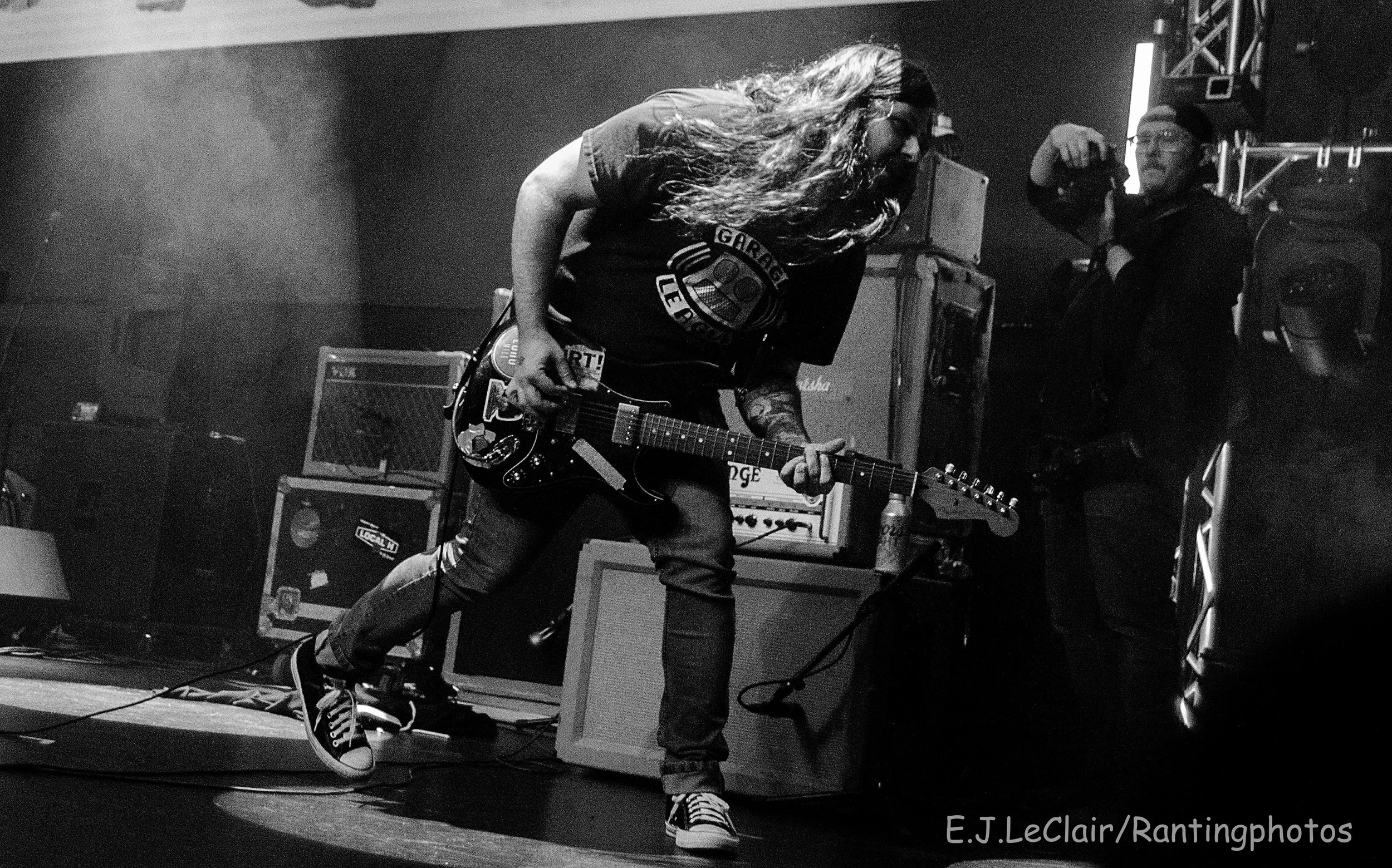 A musician with long hair, glasses, and tattoos, wearing a black t-shirt and sneakers, is playing an electric guitar on stage. There are amplifiers and sound equipment in the background. The photo is in black and white.