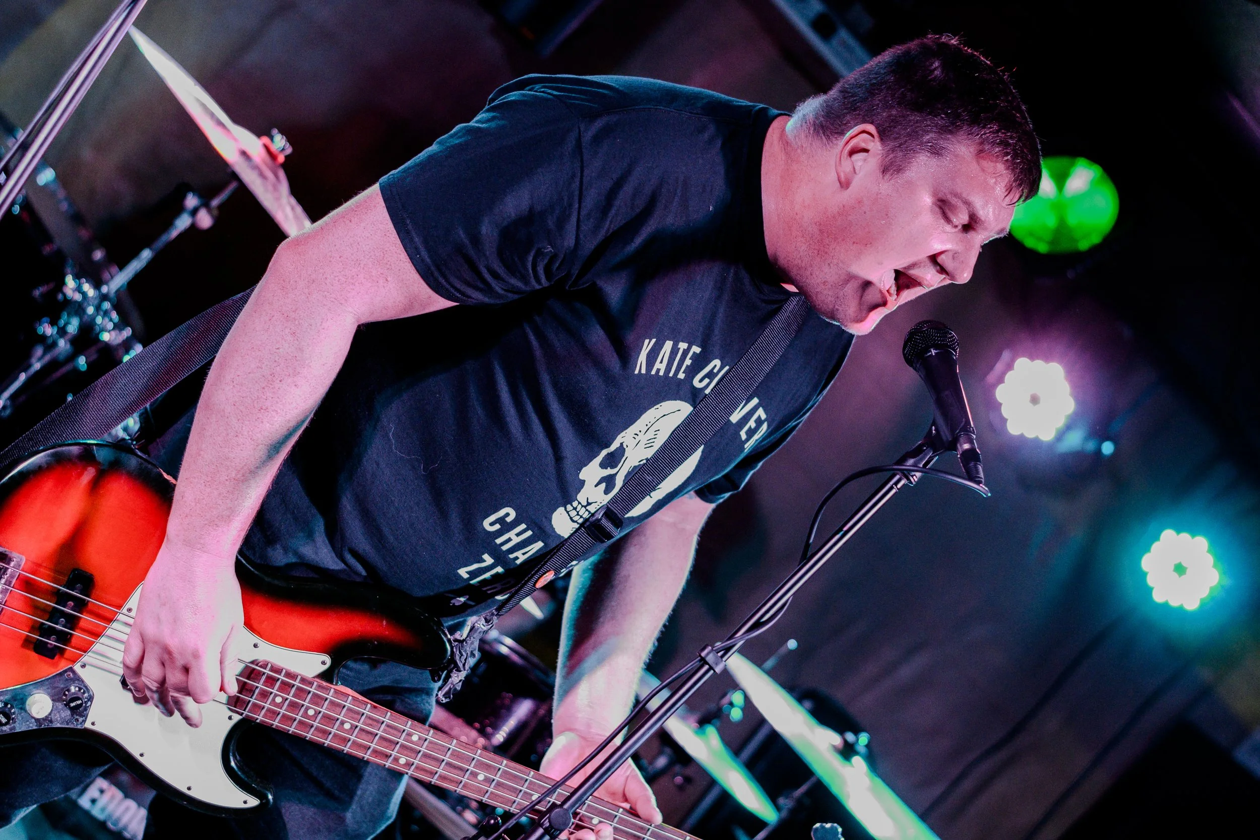 A man with short dark hair playing an orange and white electric bass guitar on stage, singing into a microphone, with colorful stage lights in the background.