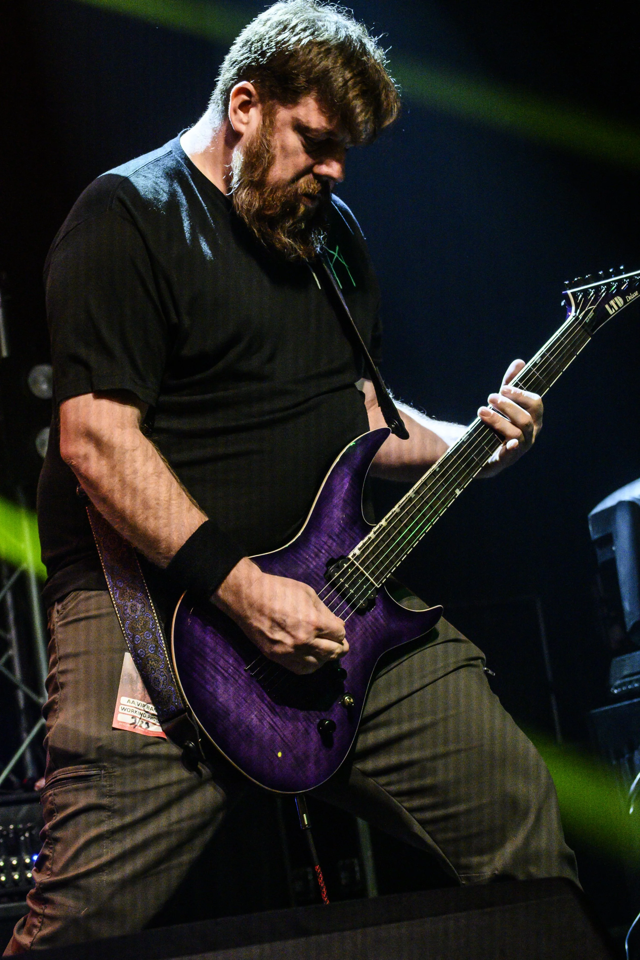 A man with a beard and glasses playing a purple electric guitar on stage, wearing a black shirt and brown striped pants.