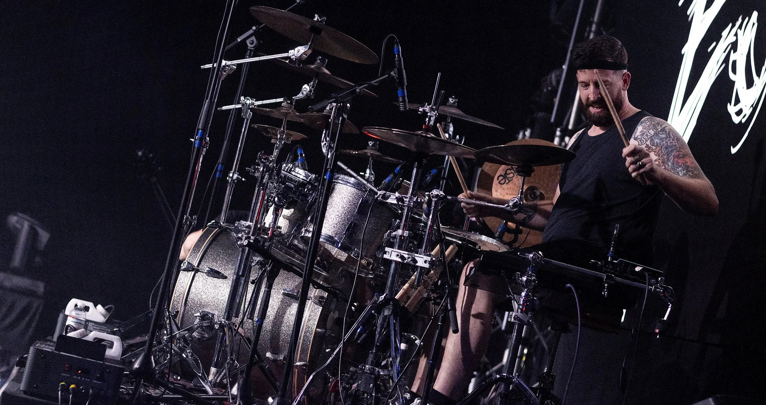 A man with tattoos on his right arm playing drums on stage with a dark background and illuminated logo or artwork behind him.