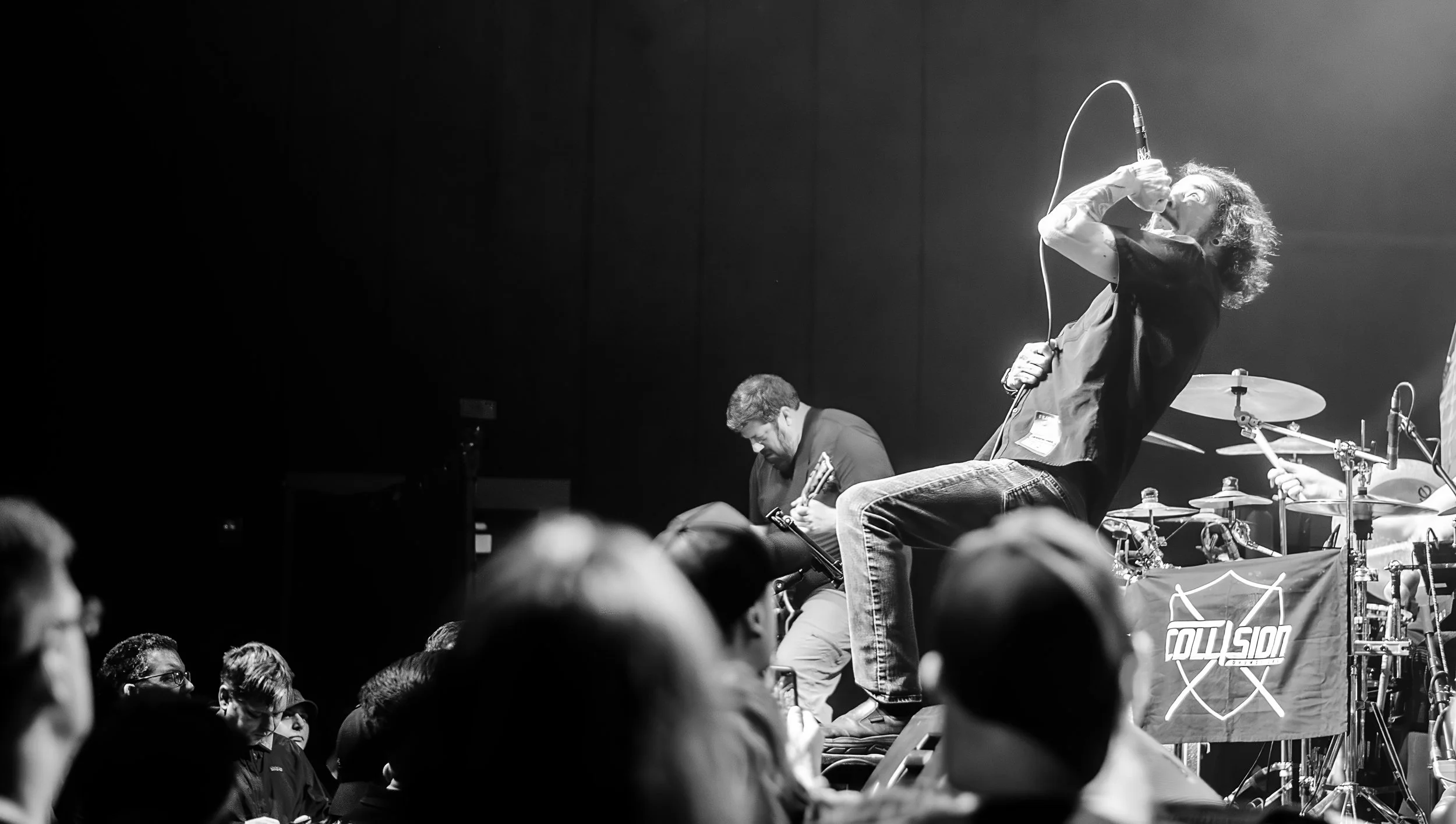 A black and white photo of a live music performance with a singer passionately singing into a microphone, and a drummer and guitarist playing on stage. The audience is visible in the foreground.