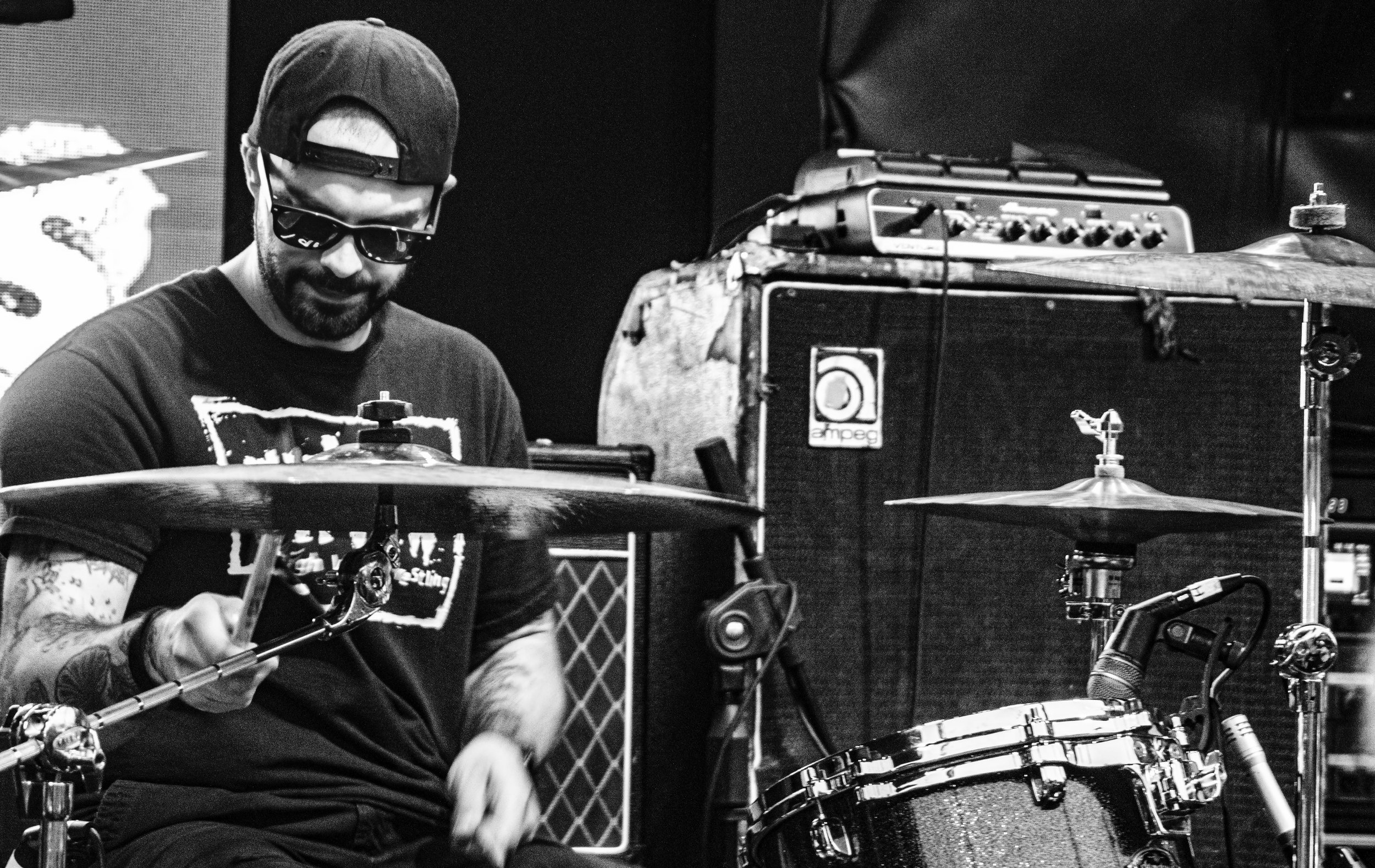 A man wearing sunglasses and a backward cap playing drums at a music venue, with musical equipment and speakers visible in the background.