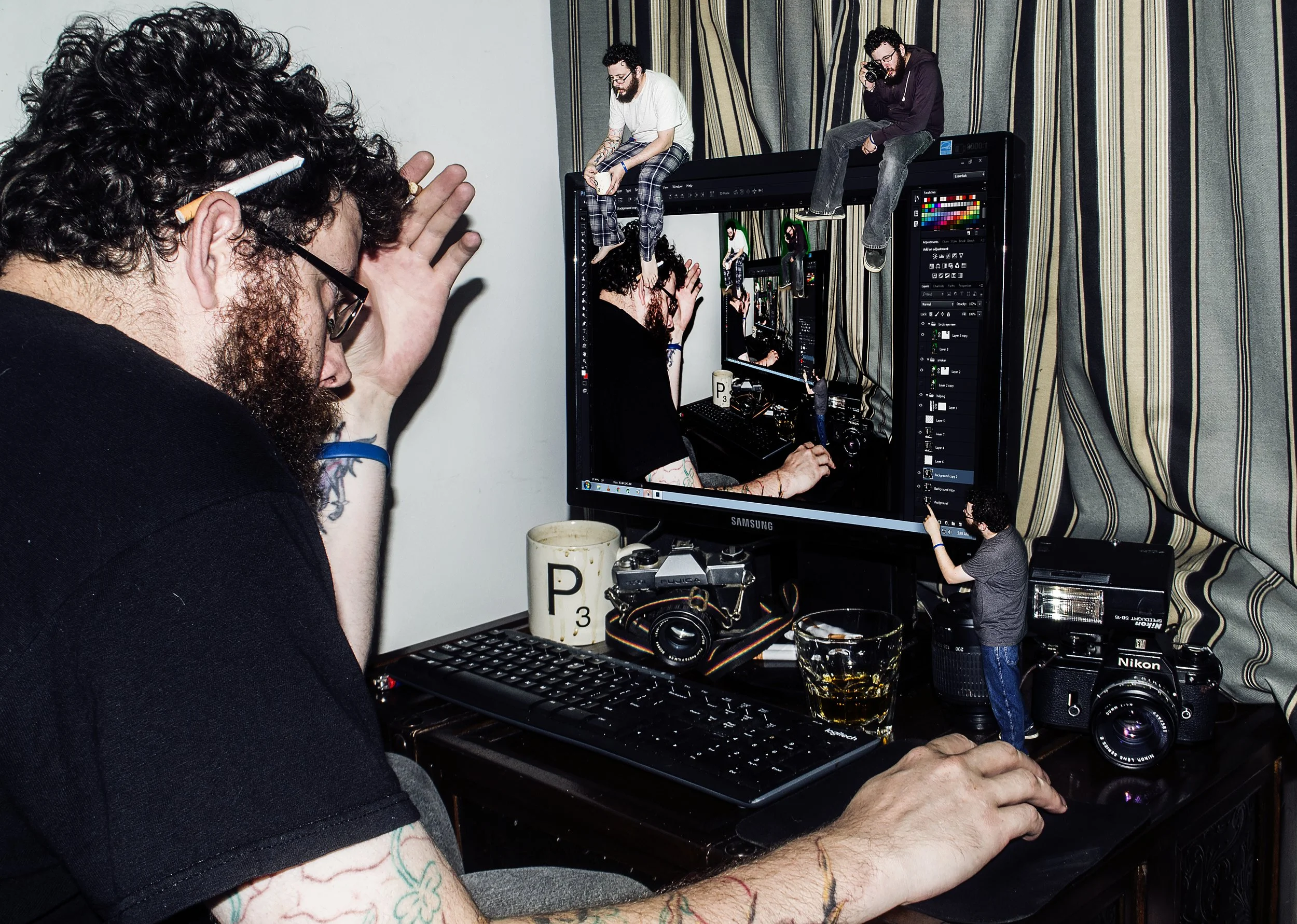 A person with tattooed arms and glasses is sitting at a desk, taking a photograph of a computer screen displaying multiple person-sized miniatures of the same scene. The scene includes two miniature figures sitting on a large computer monitor, with one holding a drink and another holding a camera, and a small child figure standing nearby holding an object.