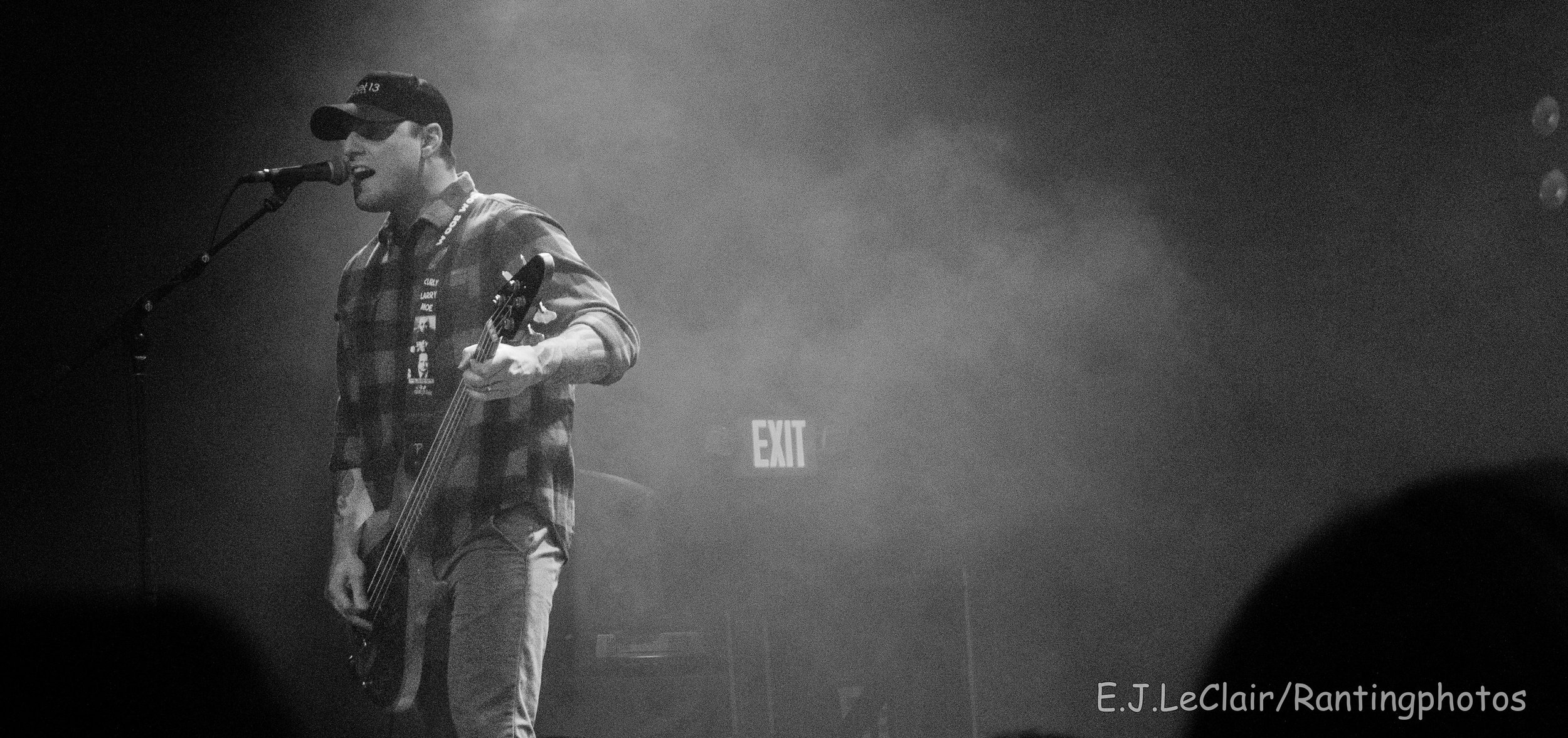 A man singing into a microphone while playing an electric bass guitar on stage. He is wearing a cap and checkered shirt, with stage fog and dim lighting. An exit sign is visible in the background.
