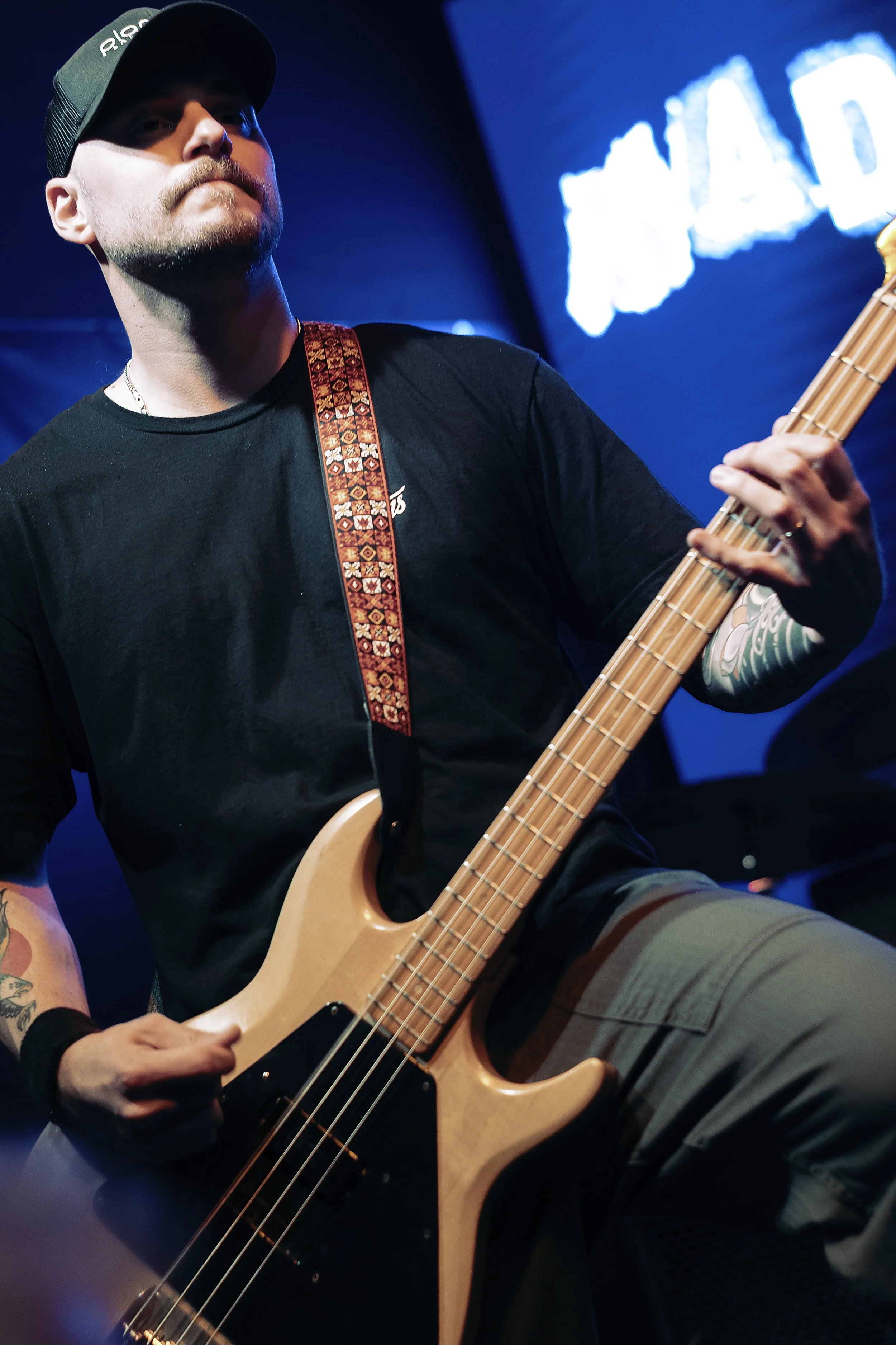 A man wearing a black shirt, a black cap, and striped shorts is playing a wooden electric bass guitar on stage. The background features a blurred sign that reads 'LOAD' with a map of Australia, illuminated with blue lighting.