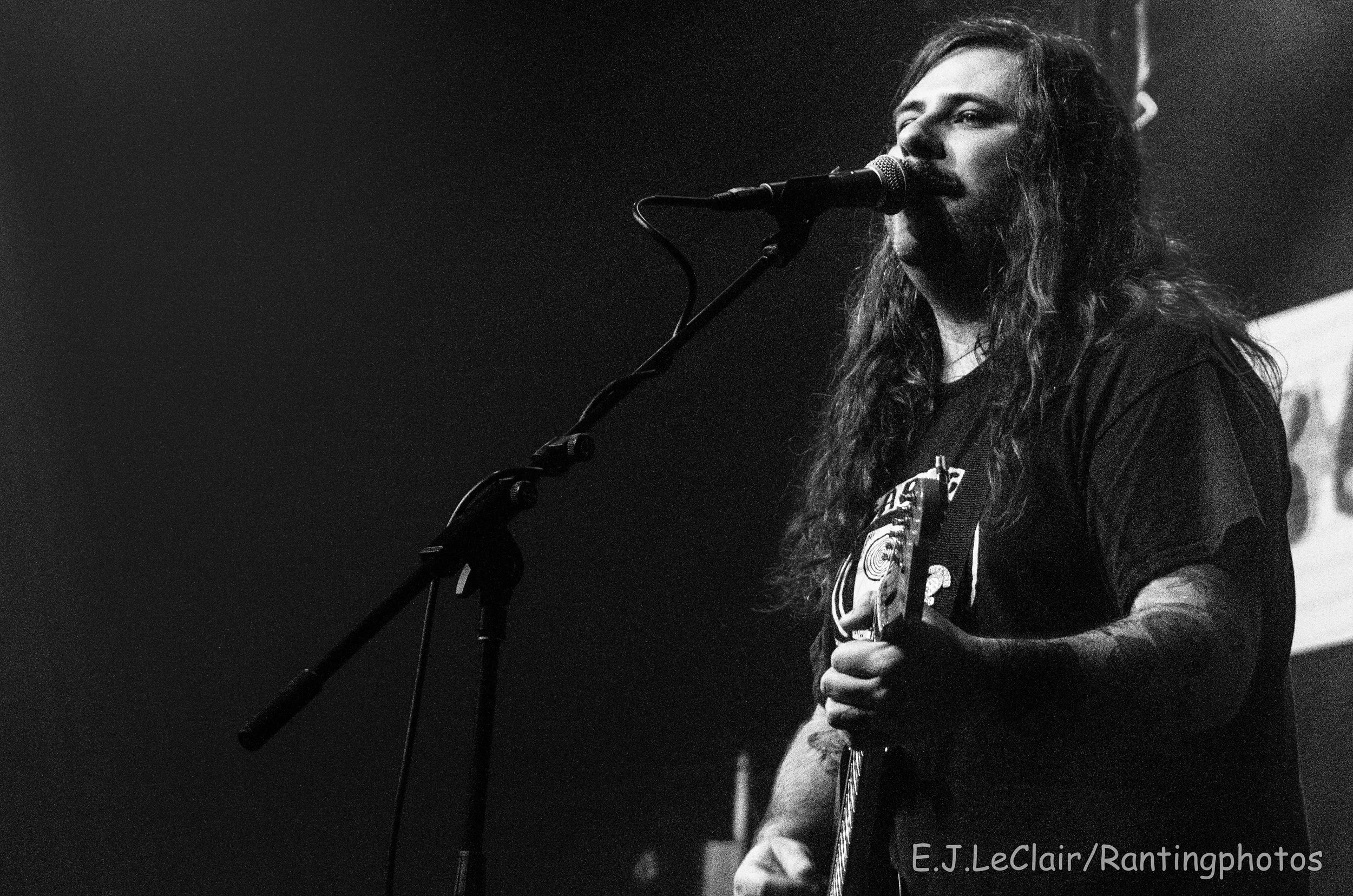 A man singing into a microphone on stage with long hair, wearing a hoodie with a graphic design, holding a guitar, in a black and white photo.
